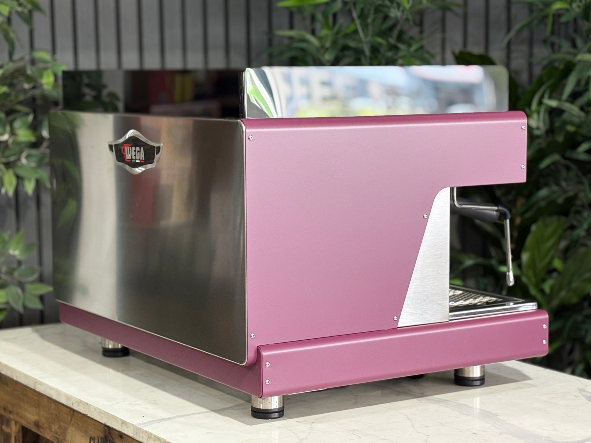 Wega Pegaso EVD 2 Group Coffee Machine Deep Pink – Used espresso coffee machine angled rear view highlighting stainless steel back panel, black acrylic top shroud, and deep pink stainless sides, placed on a marble bench with greenery.