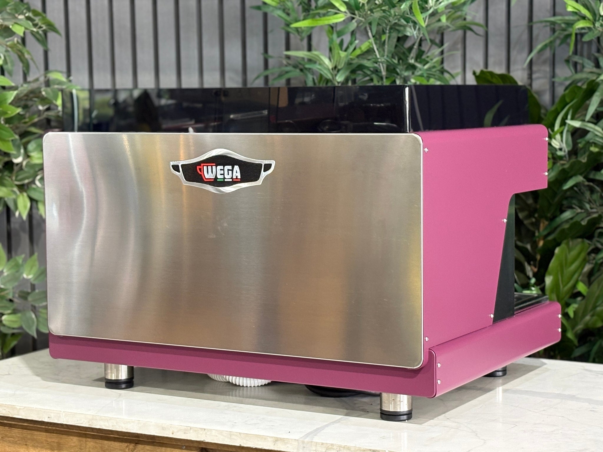 Wega Pegaso EVD 2 Group Coffee Machine Deep Pink – Used espresso coffee machine angled rear view highlighting stainless steel back panel, black acrylic top shroud, and deep pink stainless sides, placed on a marble bench with greenery.
