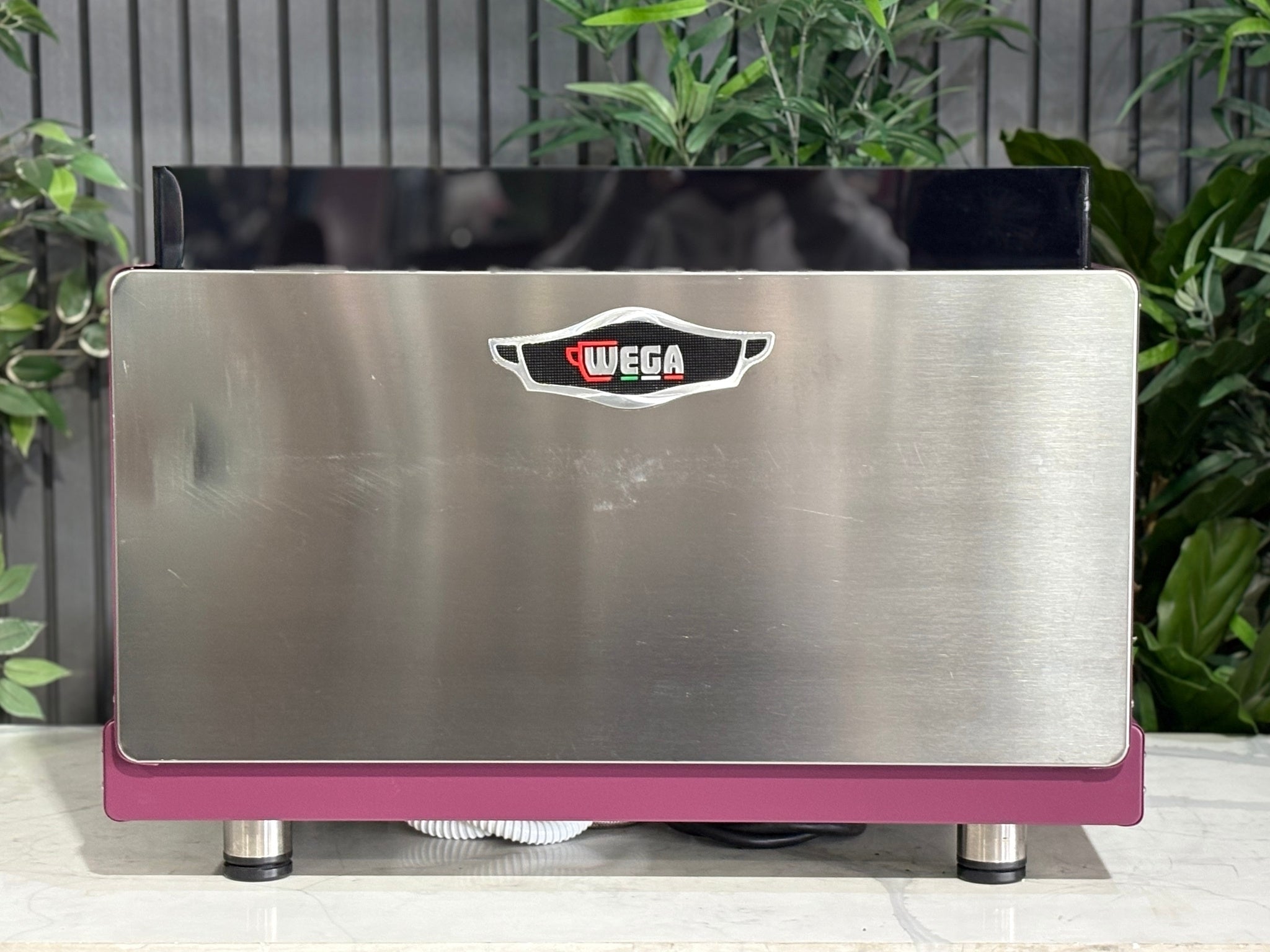 Wega Pegaso EVD 2 Group Coffee Machine Deep Pink – Used espresso coffee machine rear view showing stainless steel back panel, black acrylic top shroud, and flat deep pink stainless sides, on a marble bench with greenery.