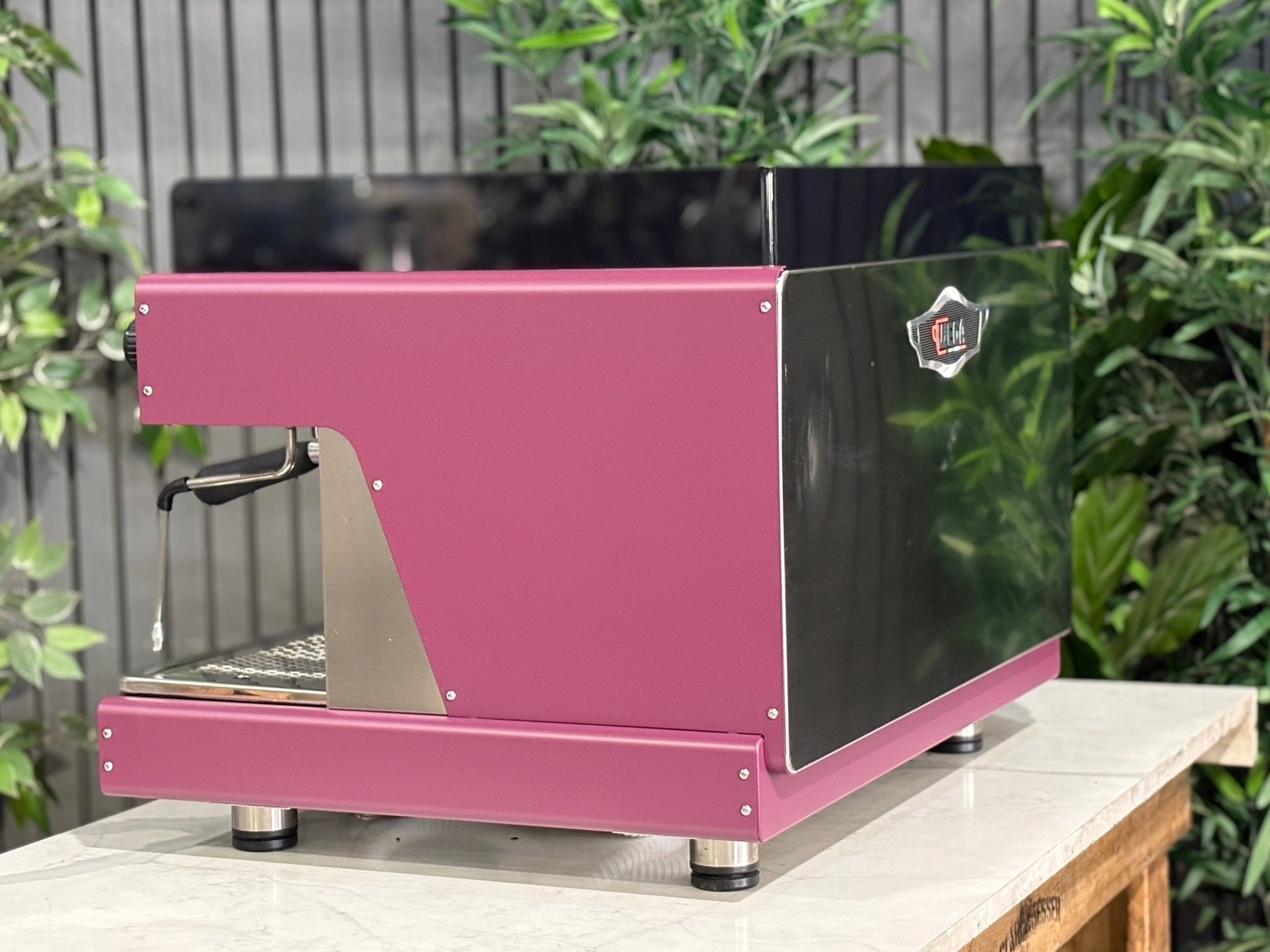 Wega Pegaso EVD 2 Group Coffee Machine Deep Pink – Used espresso coffee machine angled rear view highlighting stainless steel back panel, black acrylic top shroud, and deep pink stainless sides, placed on a marble bench with greenery.