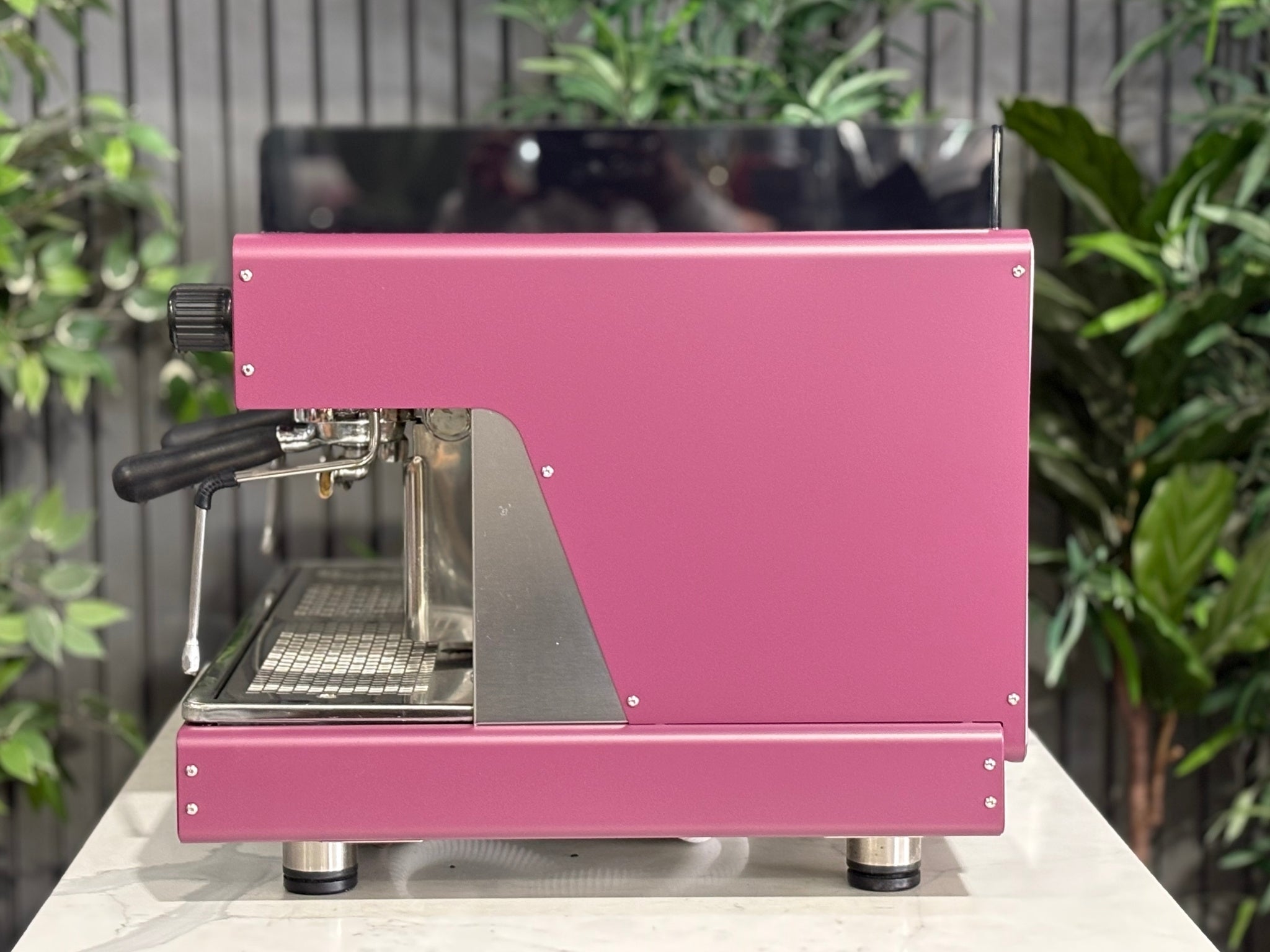 Wega Pegaso EVD 2 Group Coffee Machine Deep Pink – Used espresso coffee machine side view showing flat deep pink stainless panel, black acrylic top shroud, and black accents, on a marble bench with greenery behind.