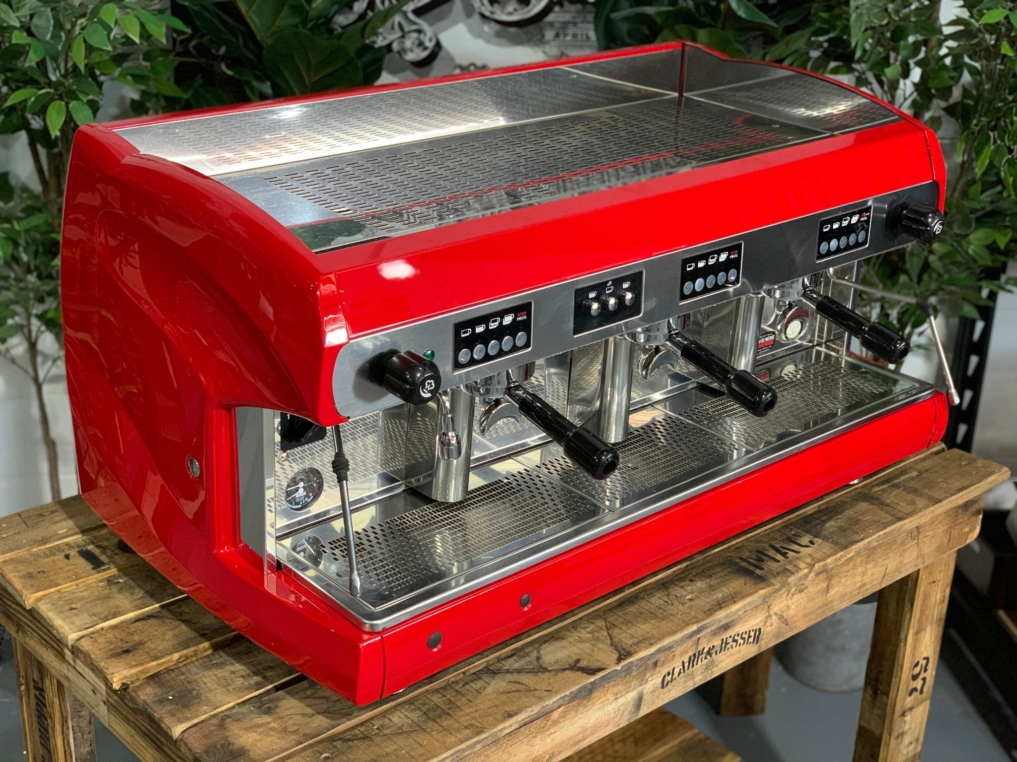 A Wega Polaris 3 Group Espresso Coffee Machine High Cup with red finish is sitting on a wooden bench. There is greenery in the background. The image is taken from the front of the machine and the machine is sitting on an angle so that you can see the front of the machine and the side.