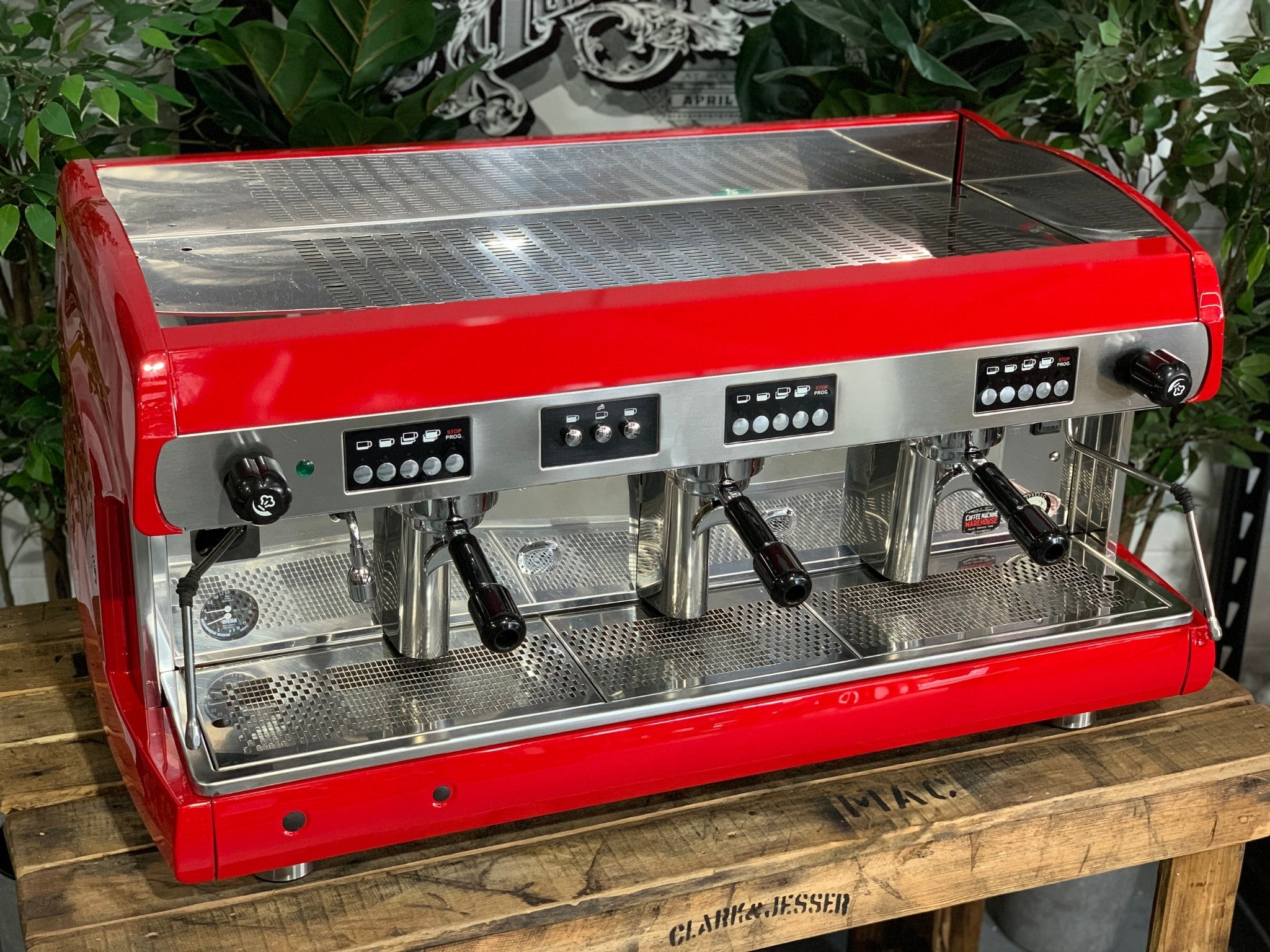 A Wega Polaris 3 Group Espresso Coffee Machine High Cup with red finish is sitting on a wooden bench. There is greenery in the background. The image is taken from the front of the machine and the machine is sitting on an angle so that you can see the front of the machine and the side.