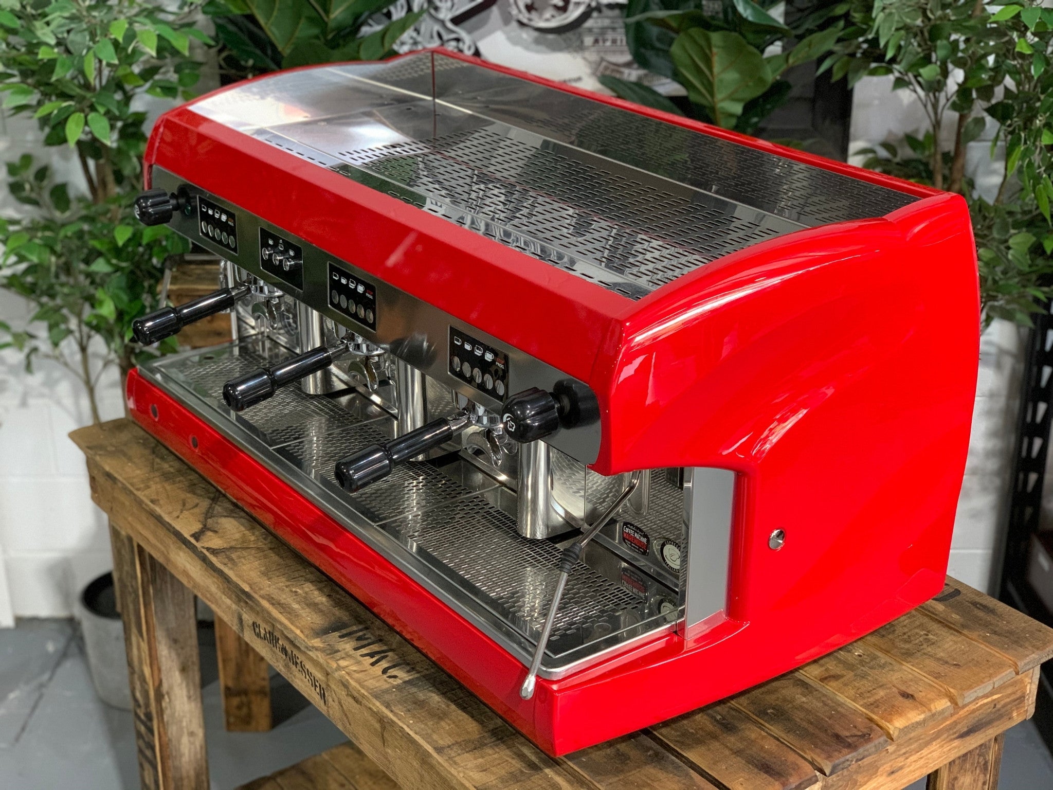 A Wega Polaris 3 Group Espresso Coffee Machine High Cup with red finish is sitting on a wooden bench. There is greenery in the background. The image is taken from the front of the machine and the machine is sitting on an angle so that you can see the front of the machine and the side.