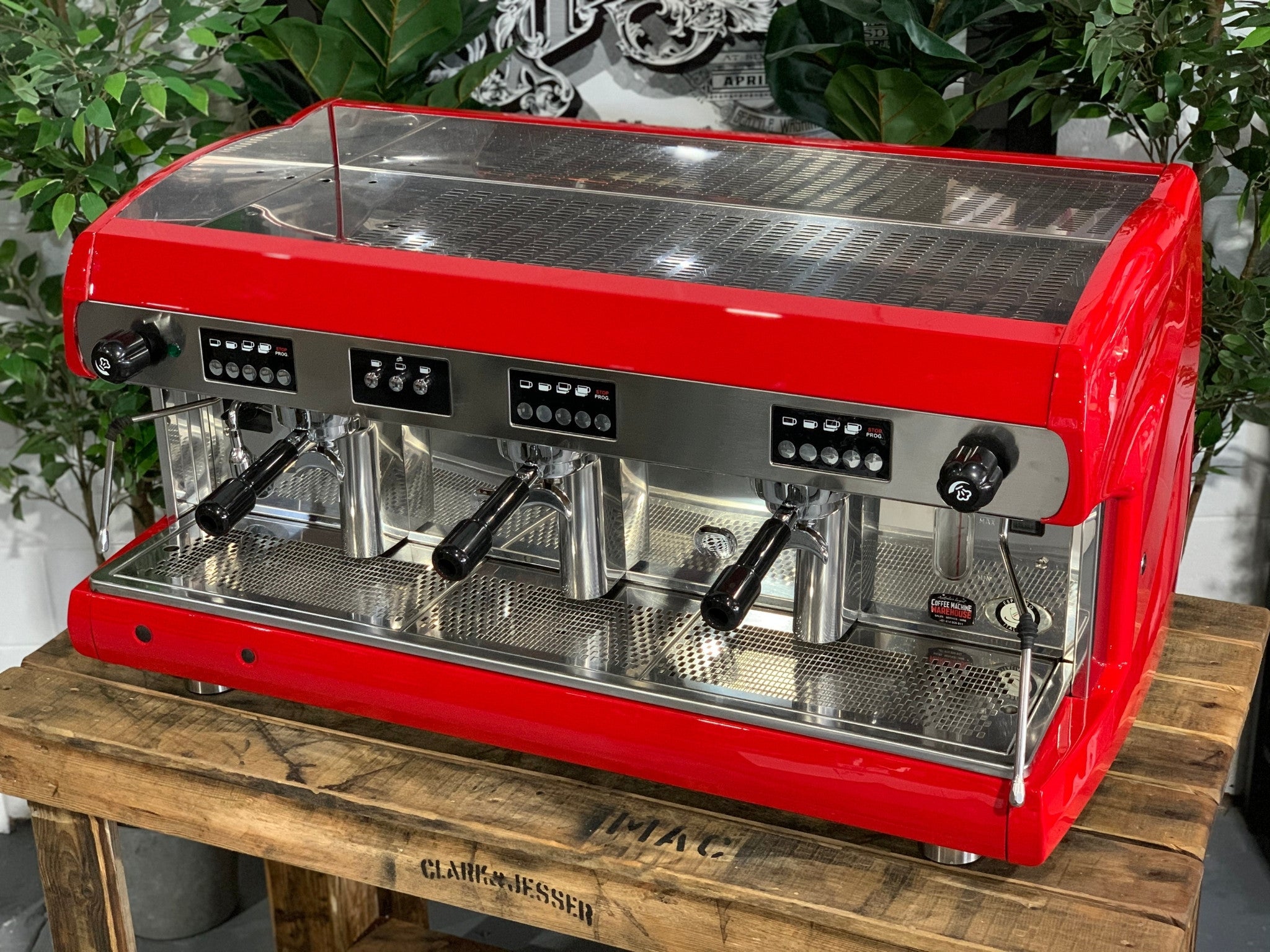 A Wega Polaris 3 Group Espresso Coffee Machine High Cup with red finish is sitting on a wooden bench. There is greenery in the background. The image is taken from the front of the machine and the machine is sitting on an angle so that you can see the front of the machine and the side.