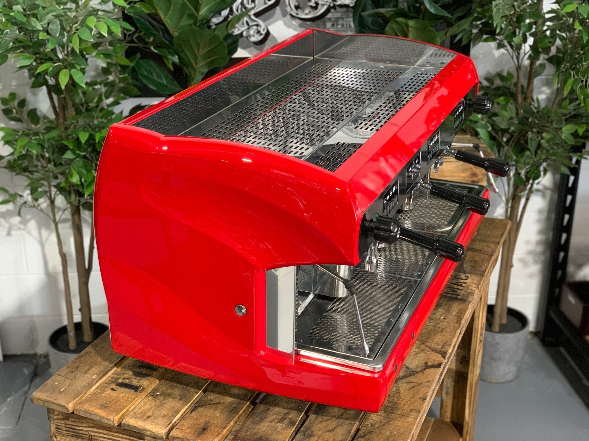 A Wega Polaris 3 Group Espresso Coffee Machine High Cup with red finish is sitting on a wooden bench. There is greenery in the background. The image is taken from the front of the machine and the machine is sitting on an angle so that you can see the front of the machine and the side.