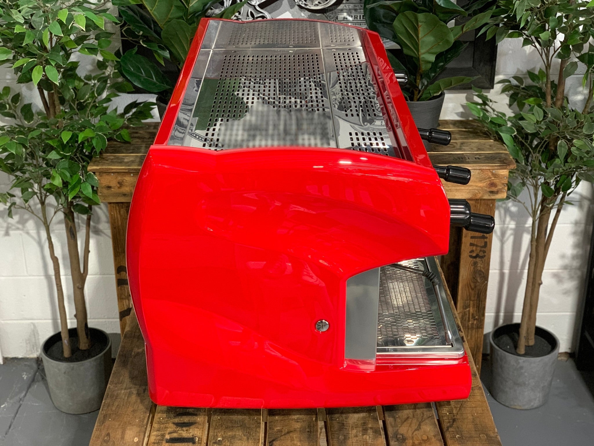 A Wega Polaris 3 Group Espresso Coffee Machine High Cup with red finish sitting on a wooden bench. There is greenery in the background. The image is taken from the side of the machine.