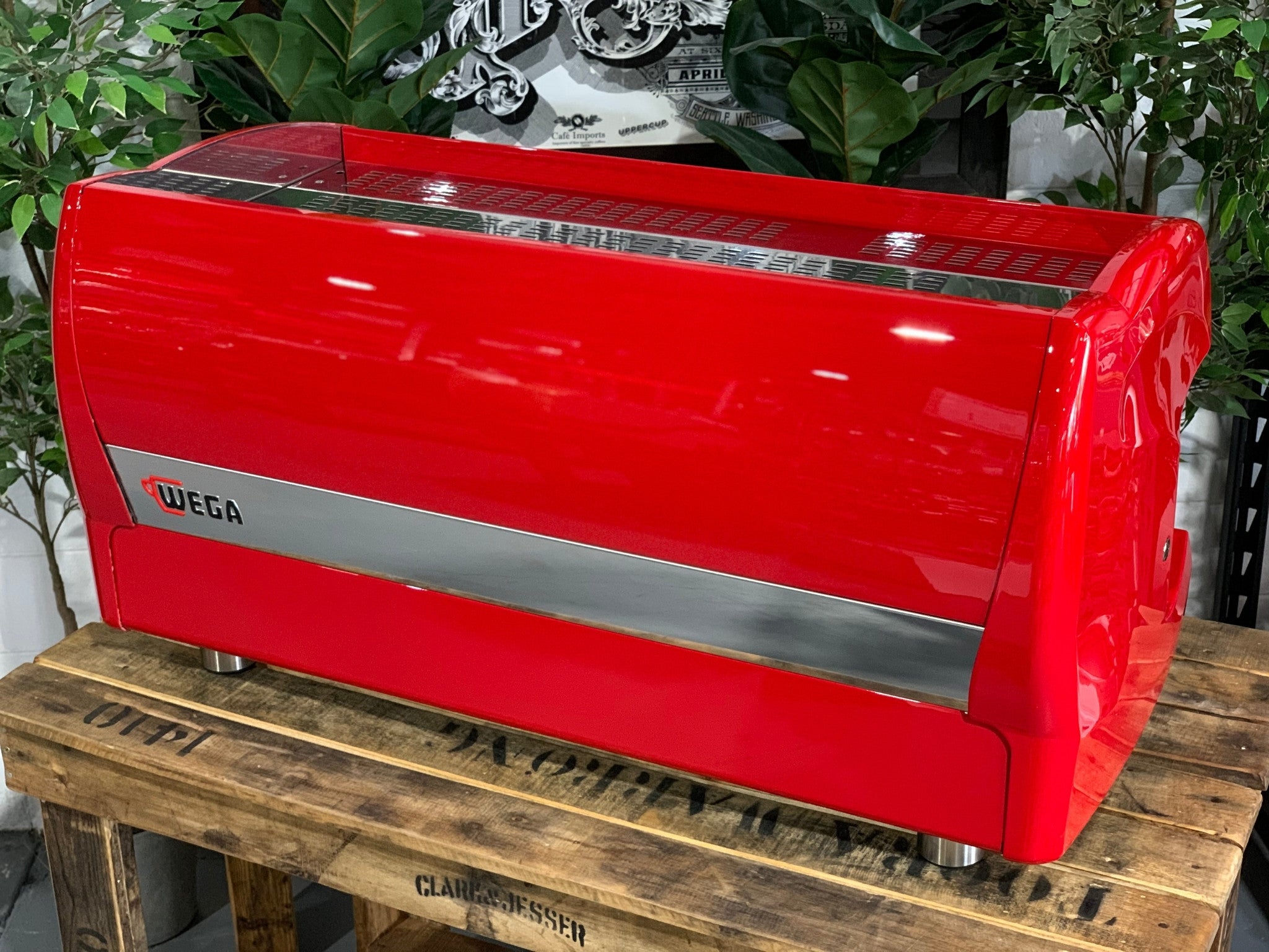 A Wega Polaris 3 Group Espresso Coffee Machine High Cup with red finish sitting on a wooden bench. There is greenery in the background. The image is taken from behind the machine and the machine is sitting on an angle so that you can see the back of the machine and the side.