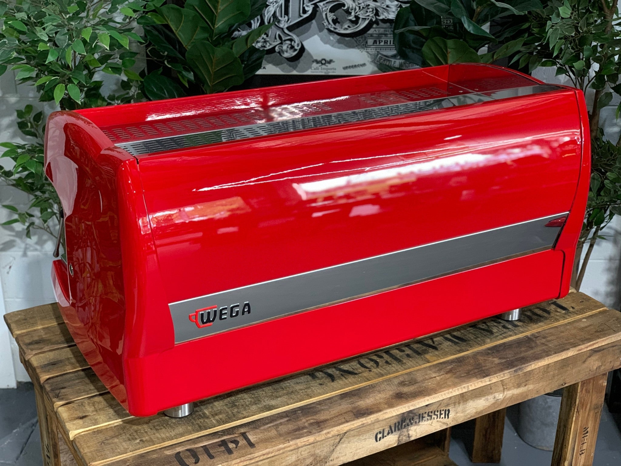 A Wega Polaris 3 Group Espresso Coffee Machine High Cup with red finishsitting on a wooden bench. There is greenery in the background. The image is taken from behind the machine and the machine is sitting on an angle so that you can see the back of the machine and the side.
The back of the machine is red, there is a thin stainless steel panel running along the back of the machine about a third down where the Wega logo is located. The logo is situated in the left hand side of that panel.