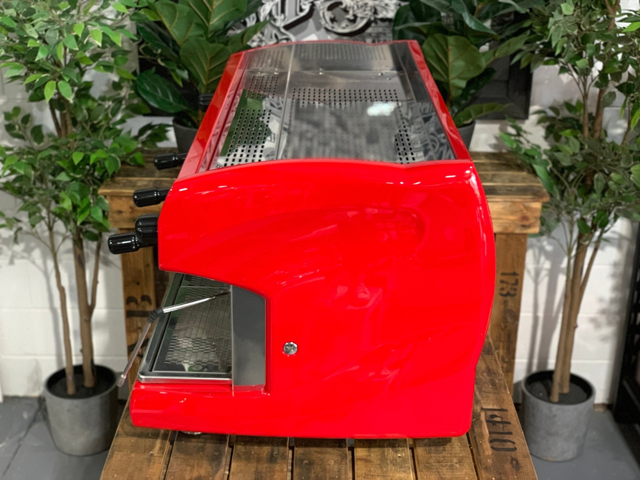A Wega Polaris 3 Group Espresso Coffee Machine High Cup with red finish sitting on a wooden bench. There is greenery in the background. The image is taken from the side of the machine.
