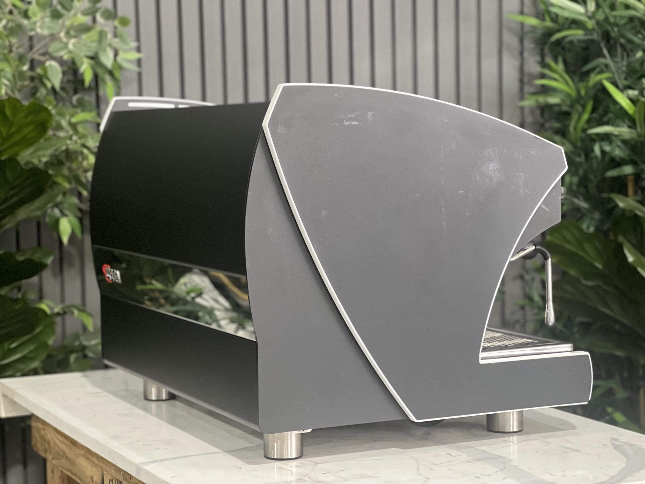 Wega Polaris Tron 2 Group Espresso Coffee Machine with Black finish sitting on a marble bench. There is greenery in the background. The image is taken from behind the machine and the machine is sitting on an angle so that you can see the back of the machine and the side.