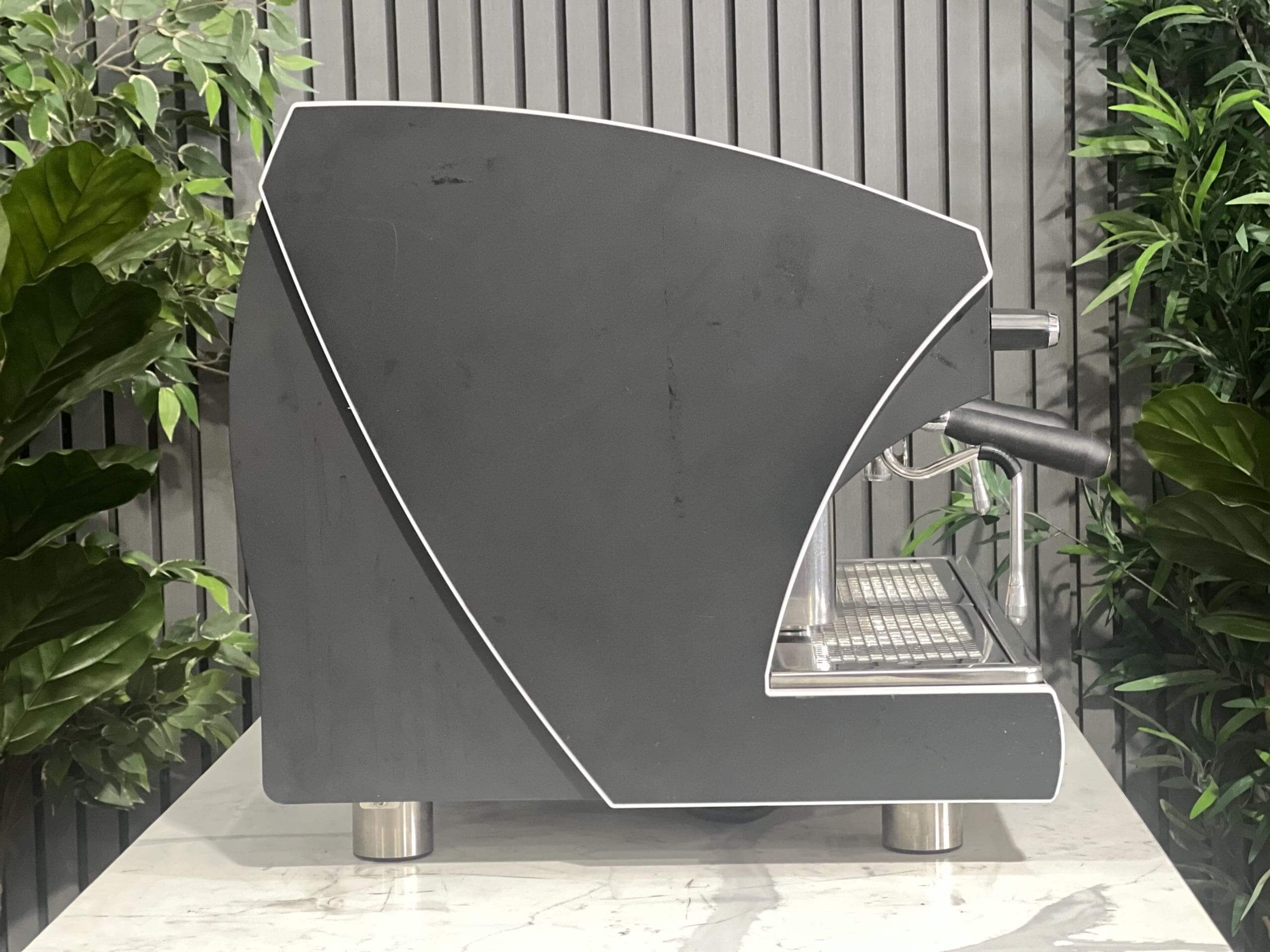 Wega Polaris Tron 2 Group Espresso Coffee Machine with Black finish sitting on a marble bench. There is greenery in the background. The image is taken from the side of the machine.