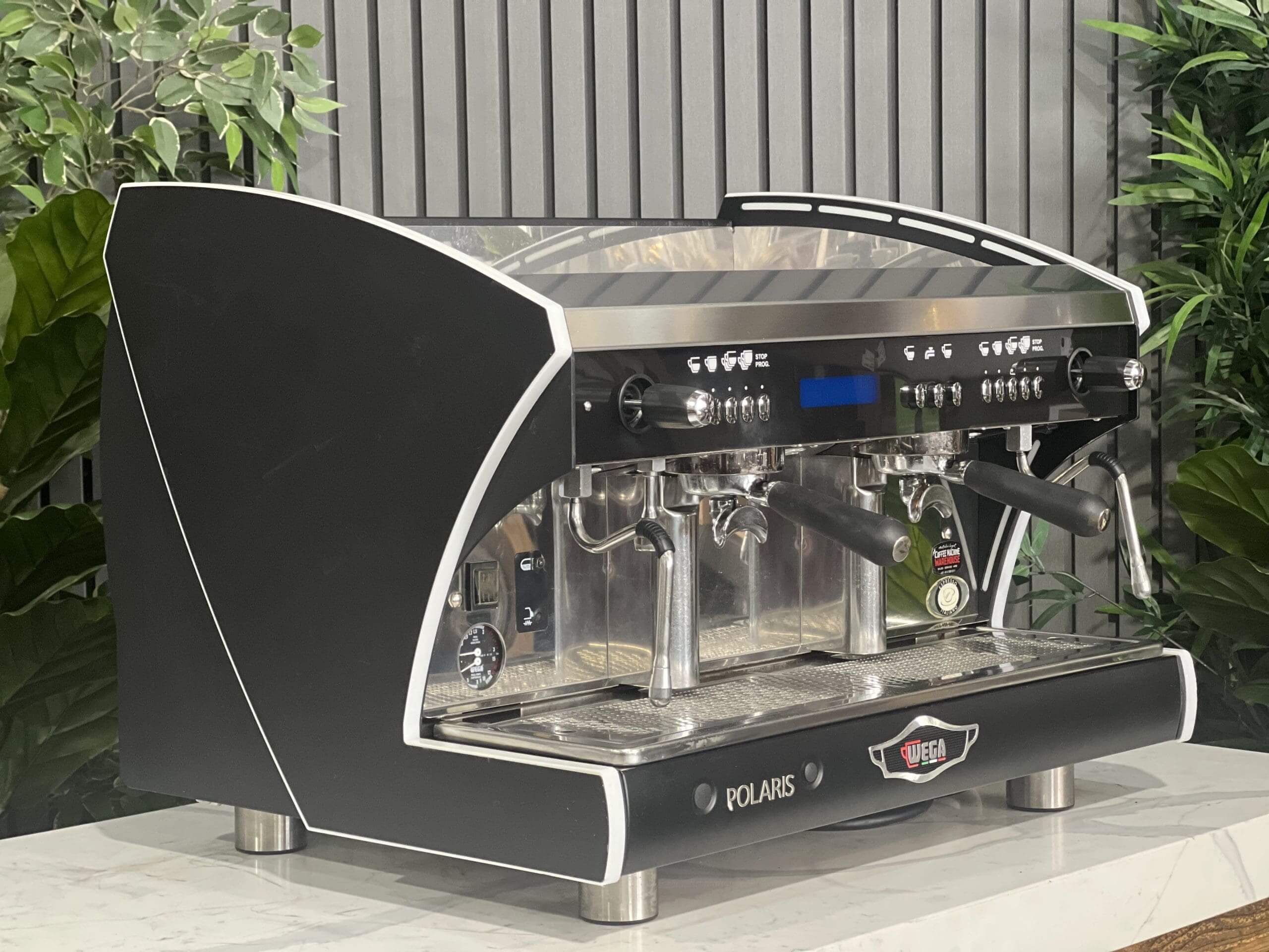 Wega Polaris Tron 2 Group Espresso Coffee Machine with Black finish  is sitting on a marble bench. There is greenery in the background. The image is taken from the front of the machine and the machine is sitting on an angle so that you can see the front of the machine and the side.