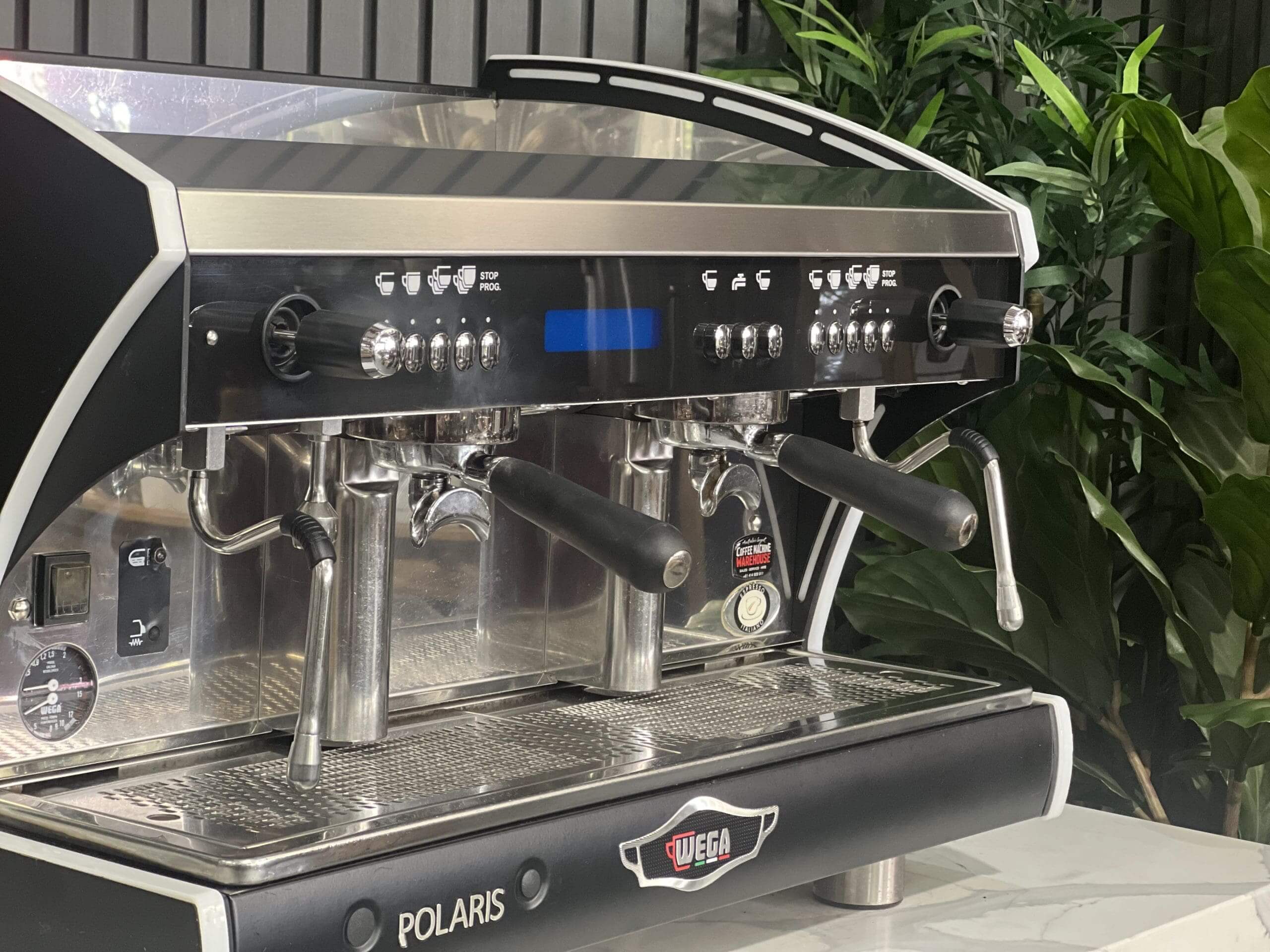 Wega Polaris Tron 2 Group Espresso Coffee Machine with Black finish is sitting on a marble bench. There is greenery in the background. The image is a close up image taken from the front of the machine and the machine is sitting on an angle so that you can see the front of the machine, the controls including buttons, group handles and logo.