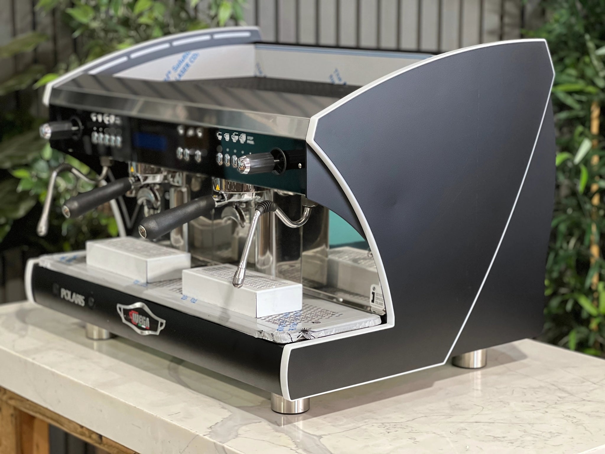 A new Wega Polaris Tron 2 Group Coffee Machine with black finish is sitting on a marble bench. There is greenery in the background. The image is taken from the front of the machine and the machine is sitting on an angle so that you can see the front of the machine and the side.