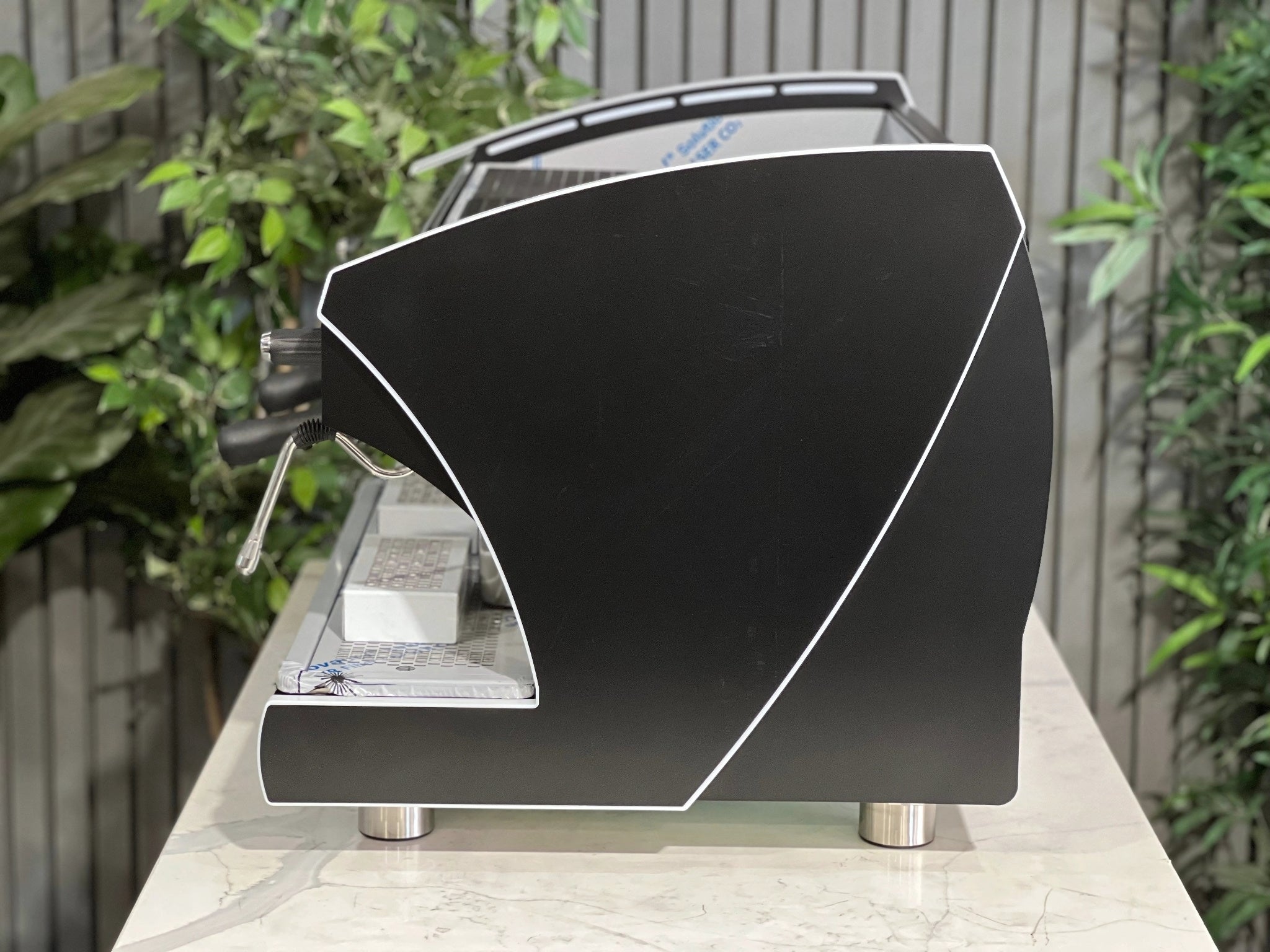 A new Wega Polaris Tron 2 Group Coffee Machine with black finish sitting on a marble bench. There is greenery in the background. The image is taken from the side of the machine.
