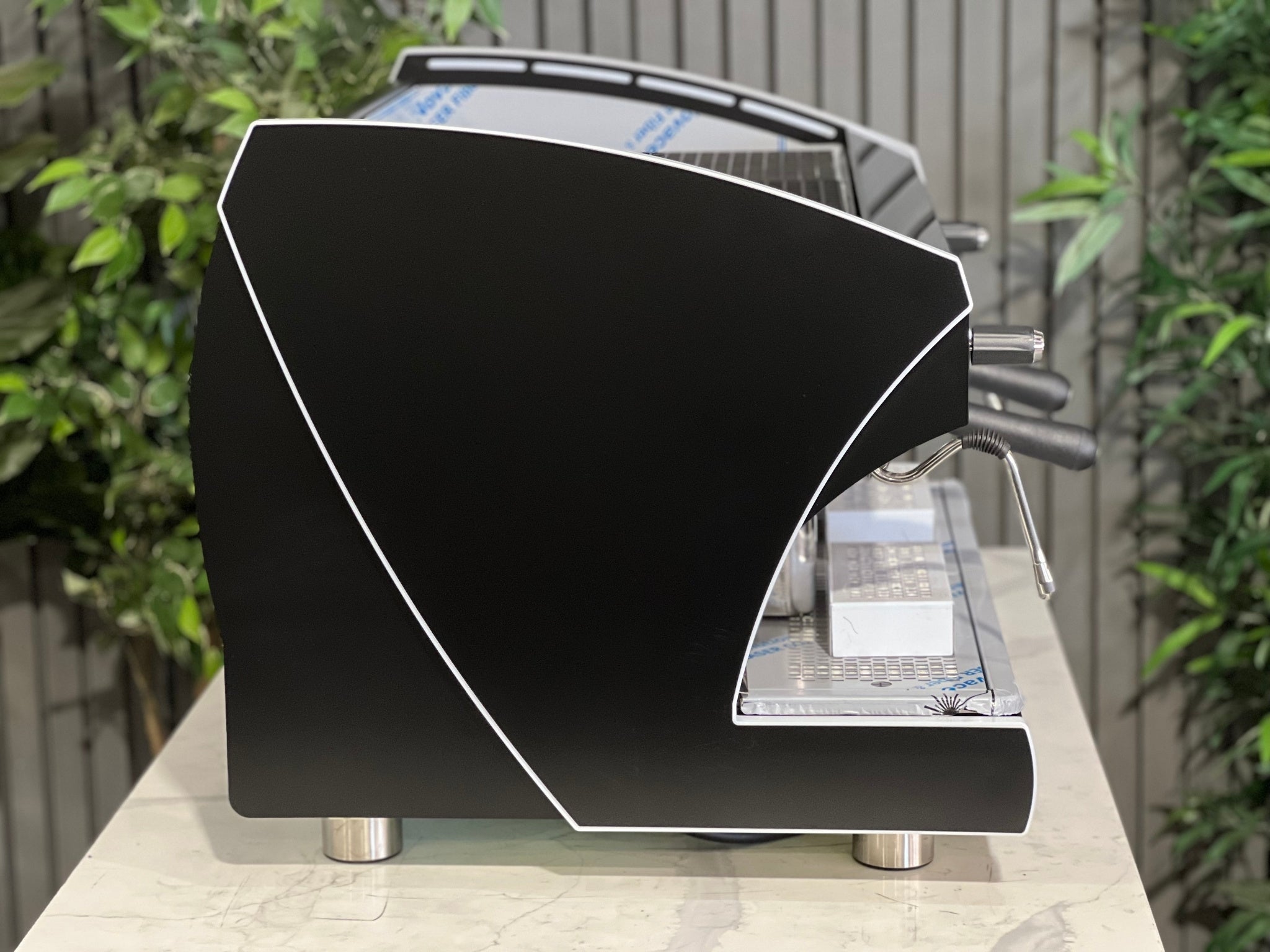 A new Wega Polaris Tron 2 Group Coffee Machine with black finish sitting on a marble bench. There is greenery in the background. The image is taken from the side of the machine.