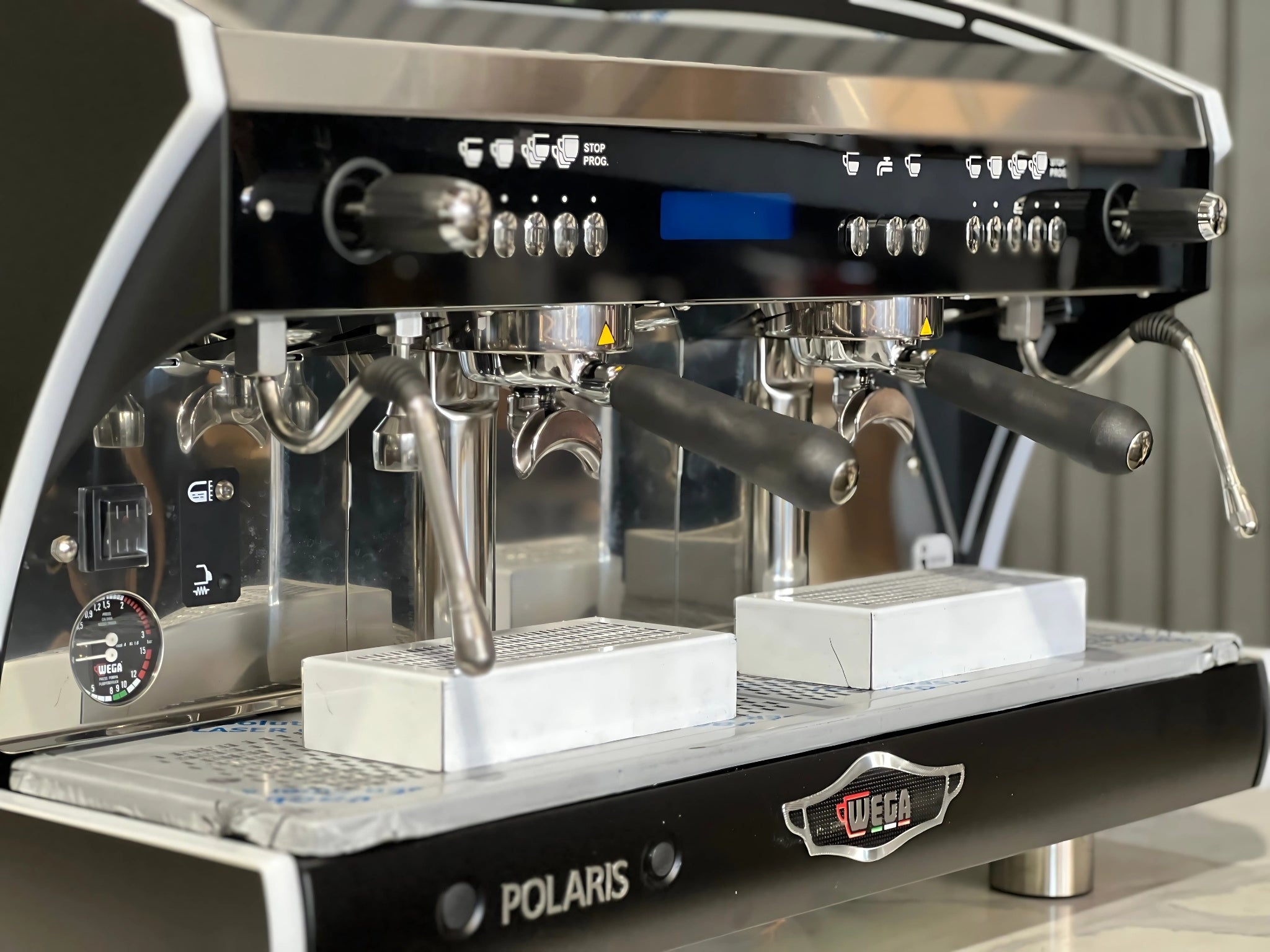 A new Wega Polaris Tron 2 Group Coffee Machine with black finish is sitting on a marble bench. There is greenery in the background. The image is a close up image taken from the front of the machine and the machine is sitting on an angle so that you can see the front of the machine, the controls including buttons, group handles and logo.