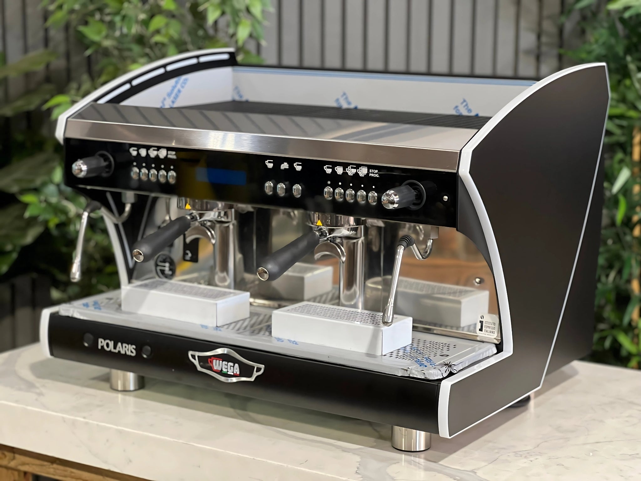 A new Wega Polaris Tron 2 Group Coffee Machine with black finish is sitting on a marble bench. There is greenery in the background. The image is taken from the front of the machine and the machine is sitting on an angle so that you can see the front of the machine and the side.