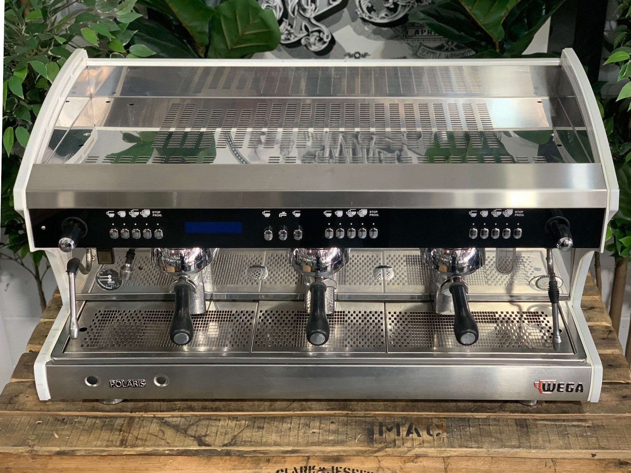 A Wega Polaris Tron 3 Group Espresso Coffee Machine with white finish sitting on a wooden bench. There is greenery in the background. The image is taken from the front of the machine but a higher angle so that you can see the front of the machine and also the top of the machine.