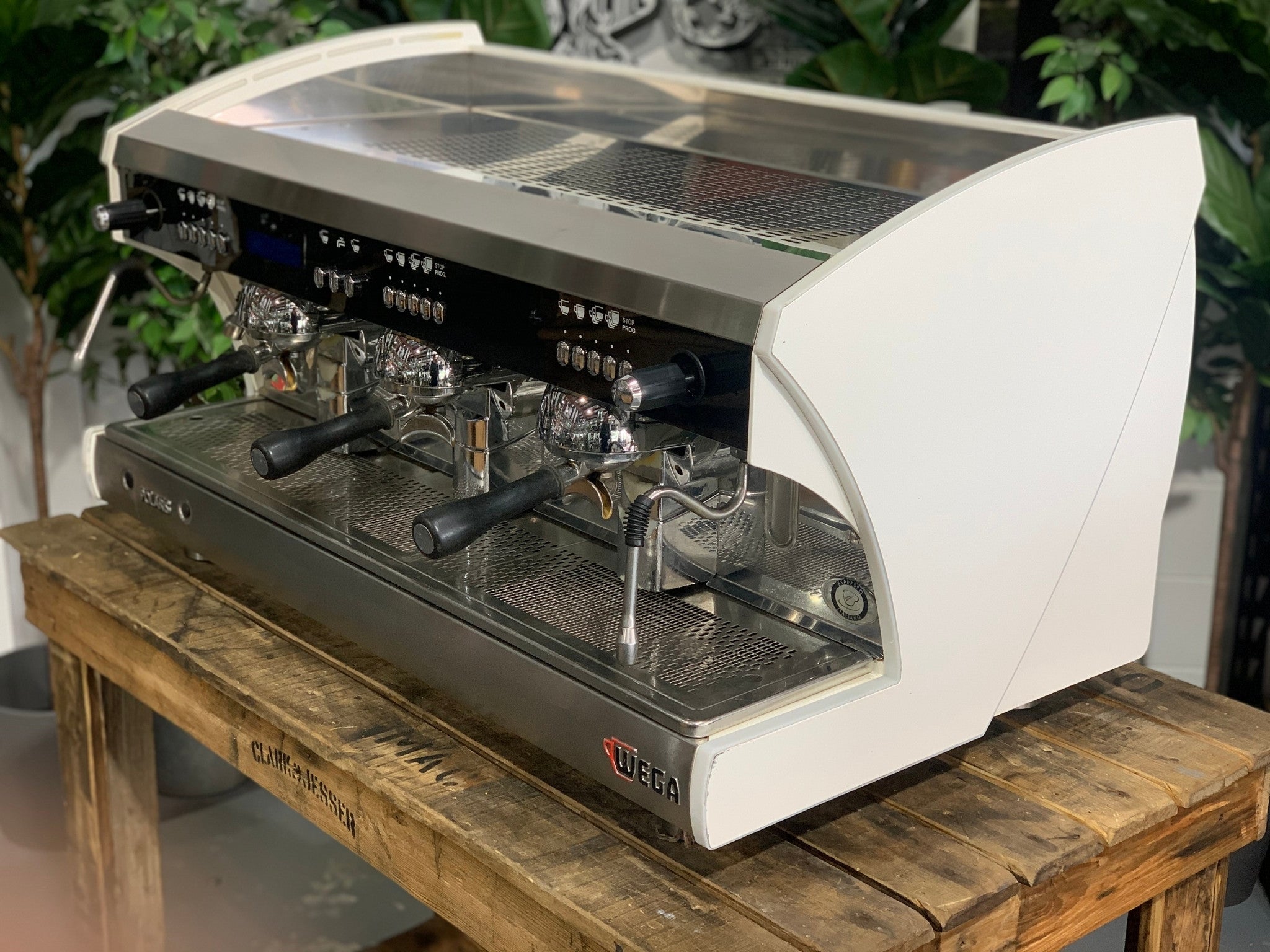 A Wega Polaris Tron 3 Group Espresso Coffee Machine with white finish  is sitting on a wooden bench. There is greenery in the background. The image is taken from the front of the machine and the machine is sitting on an angle so that you can see the front of the machine and the side.