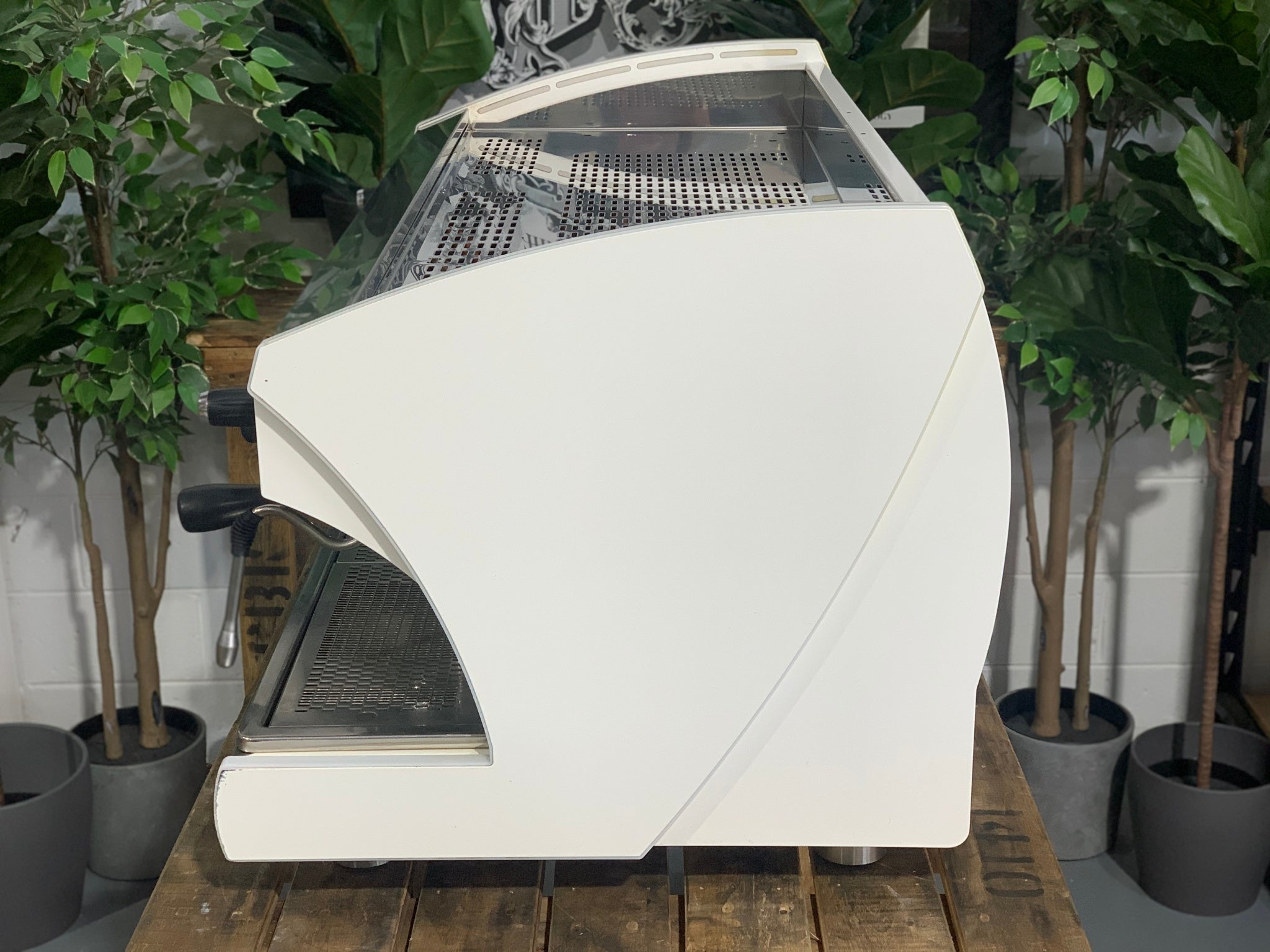 A Wega Polaris Tron 3 Group Espresso Coffee Machine with white finish sitting on a wooden bench. There is greenery in the background. The image is taken from the side of the machine.