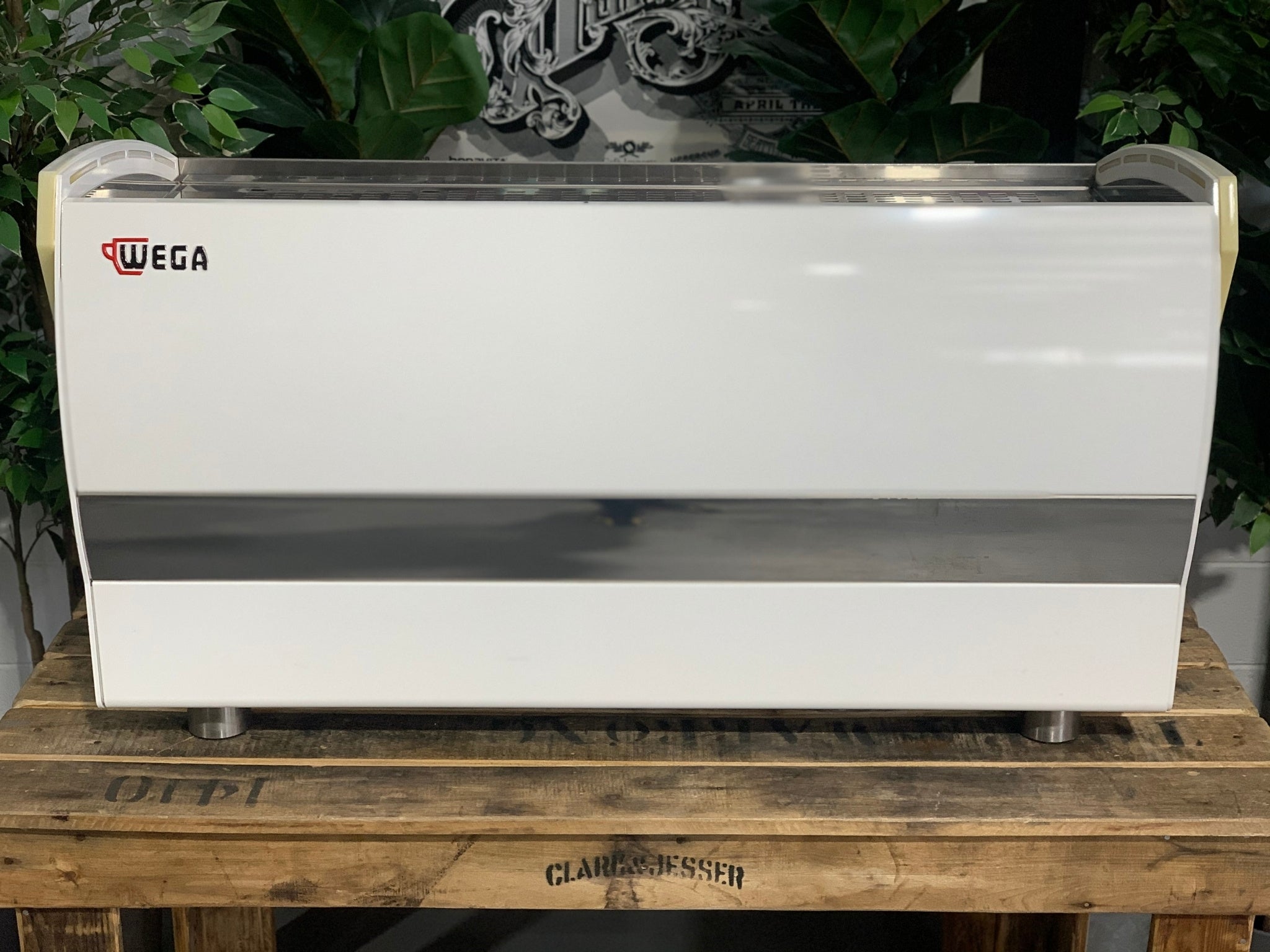 A Wega Polaris Tron 3 Group Espresso Coffee Machine with white finish is sitting on a wooden bench. There is greenery in the background. The image is taken from behind the machine so the back is visible. The Wega logo is situated in the top left hand corner of the back panel.