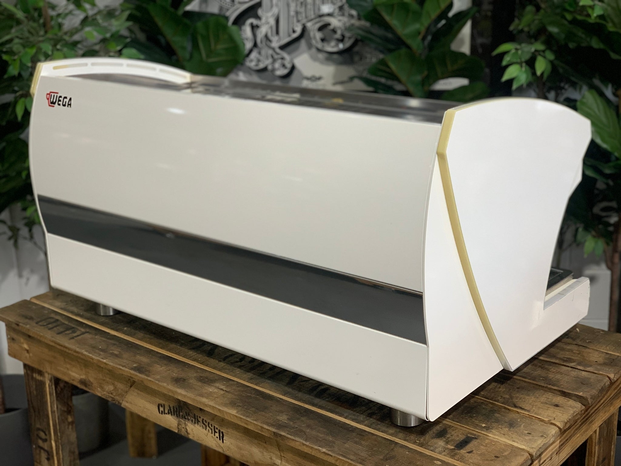 A Wega Polaris Tron 3 Group Espresso Coffee Machine with white finish sitting on a wooden bench. There is greenery in the background. The image is taken from behind the machine and the machine is sitting on an angle so that you can see the back of the machine and the side.