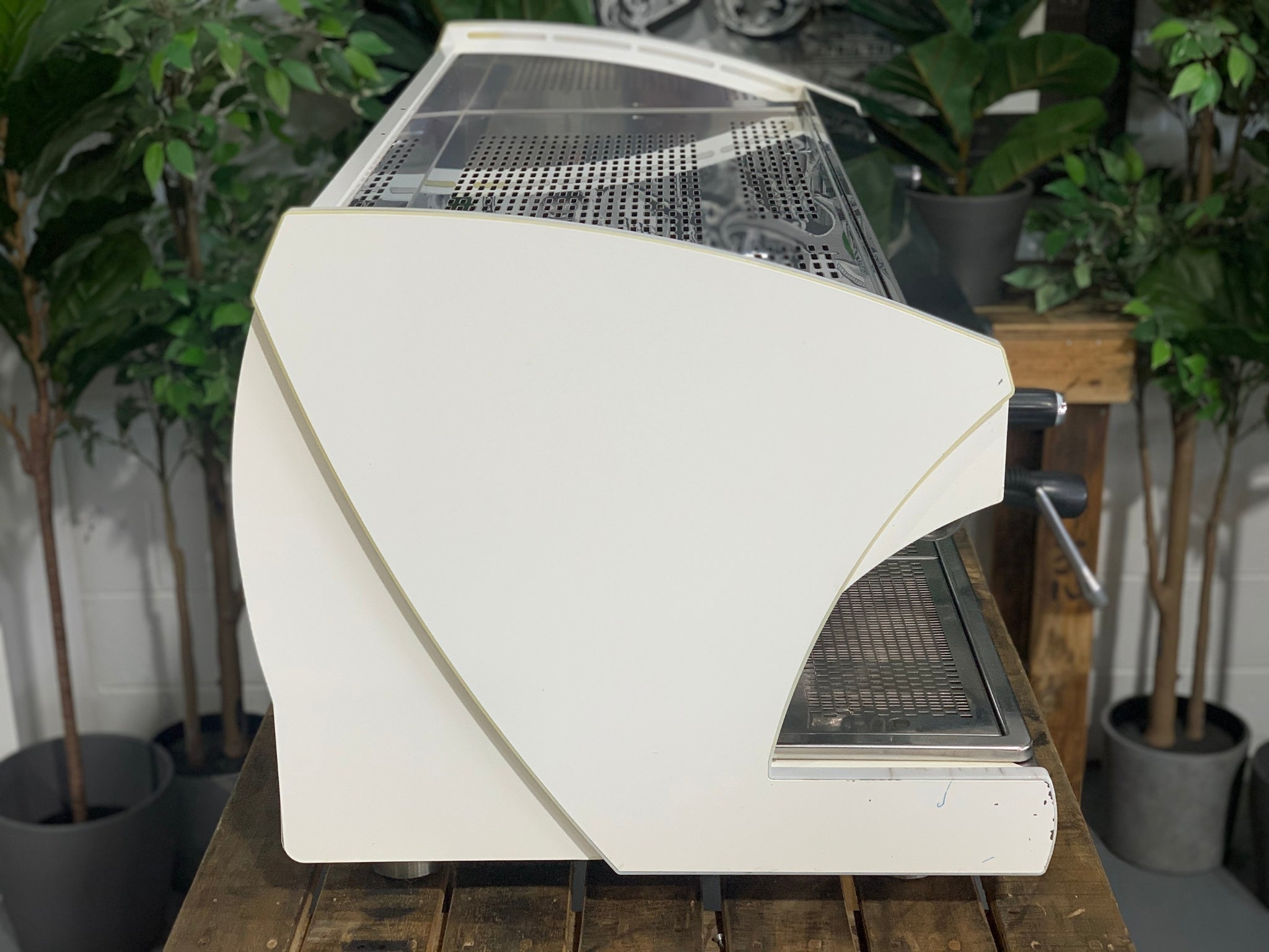 A Wega Polaris Tron 3 Group Espresso Coffee Machine with white finish sitting on a wooden bench. There is greenery in the background. The image is taken from the side of the machine.