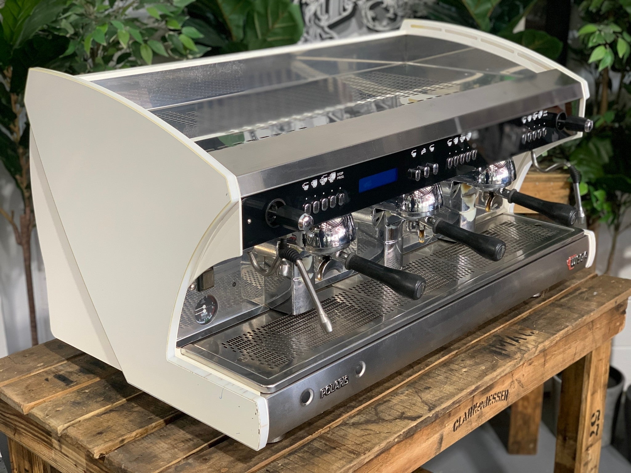 A Wega Polaris Tron 3 Group Espresso Coffee Machine with white finish is sitting on a wooden bench. There is greenery in the background. The image is taken from the front of the machine and the machine is sitting on an angle so that you can see the front of the machine and the side.