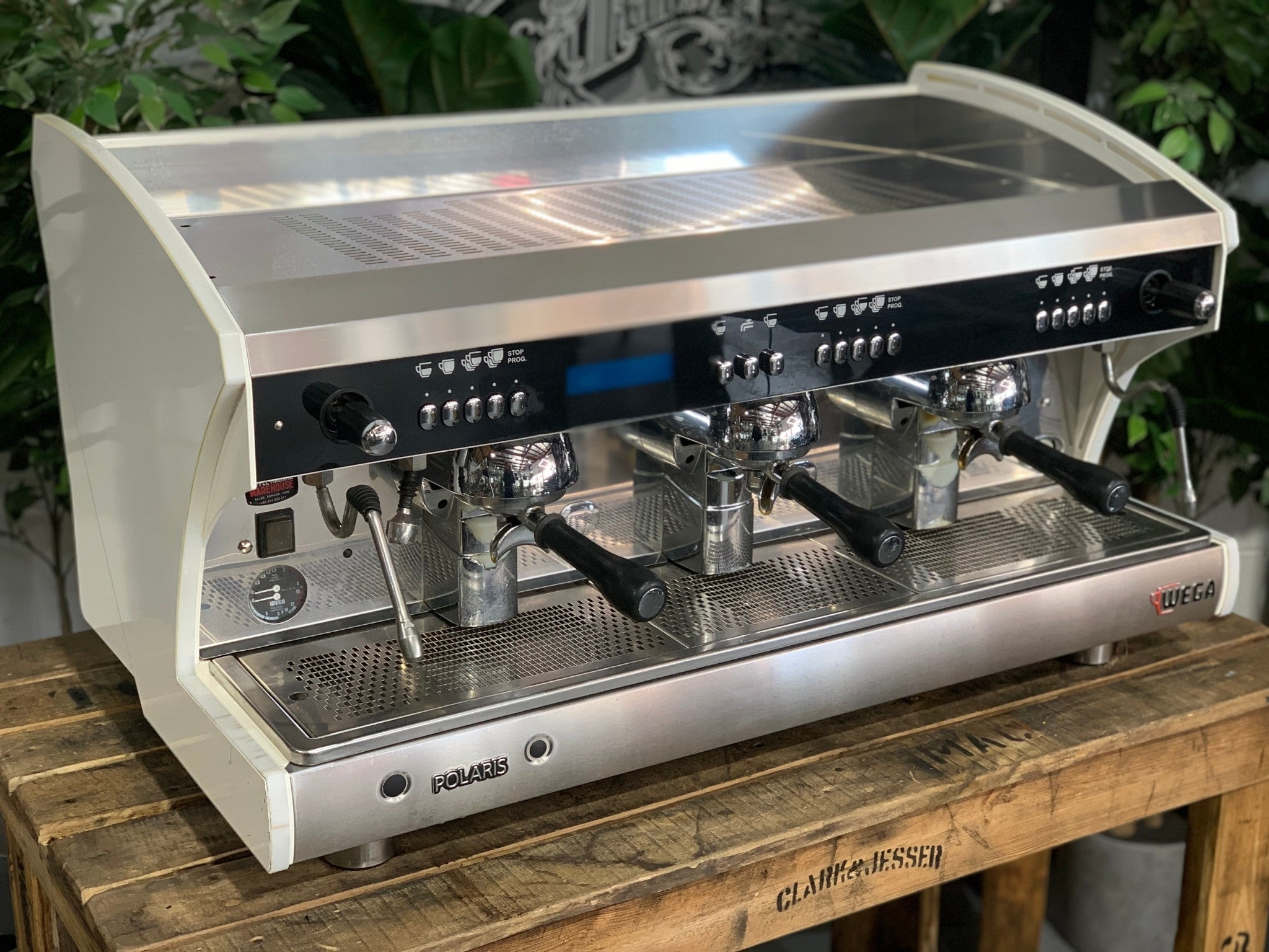 A Wega Polaris Tron 3 Group Espresso Coffee Machine with white finish is sitting on a wooden bench. There is greenery in the background. The image is taken from the front of the machine and the machine is sitting on an angle so that you can see the front of the machine and the side.