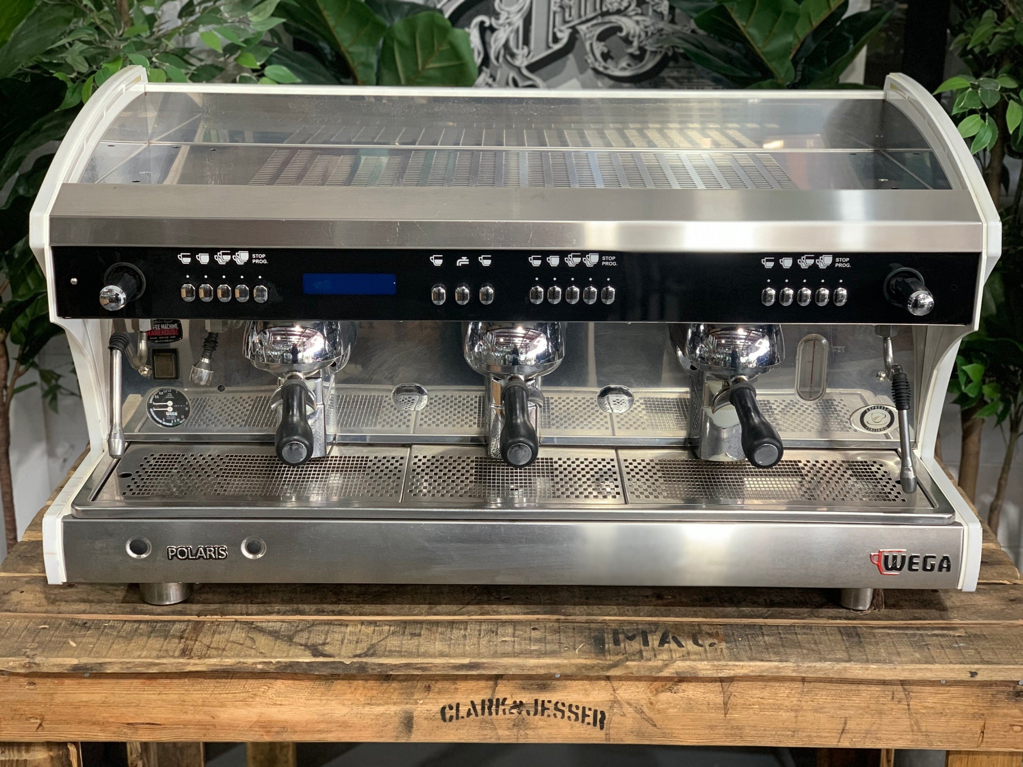 A Wega Polaris Tron 3 Group Espresso Coffee Machine with white finish  sitting on a wooden bench. There is greenery in the background. The image is taken from the front of the machine so that you can see the front of the machine, control panel, buttons and group heads.