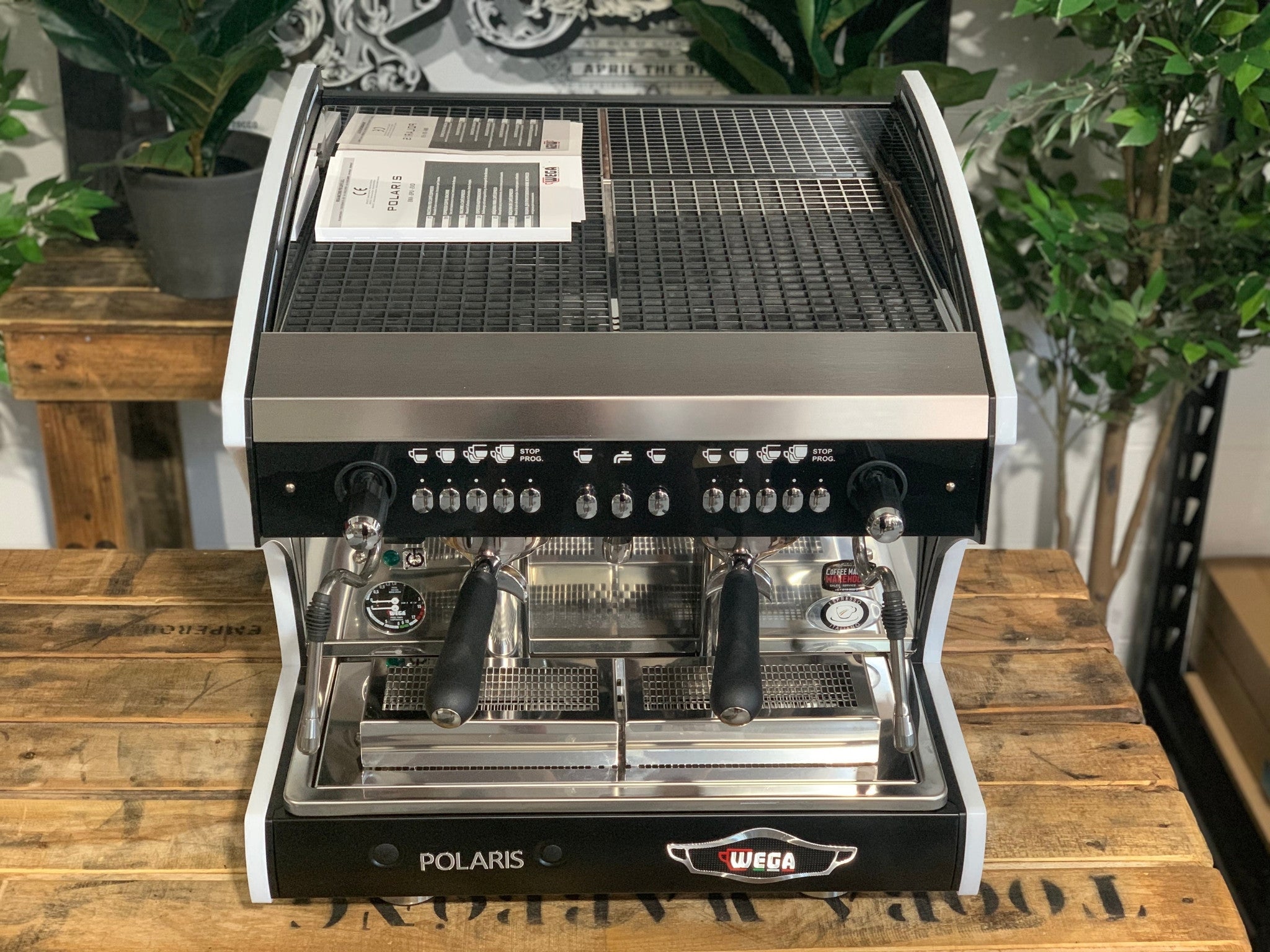 A new Wega Polaris Tron Compact 2 Group Espresso Coffee Machine with black finish sitting on a wooden bench. There is greenery in the background. The image is taken from the front of the machine but a higher angle so that you can see the front of the machine and also the top of the machine.
