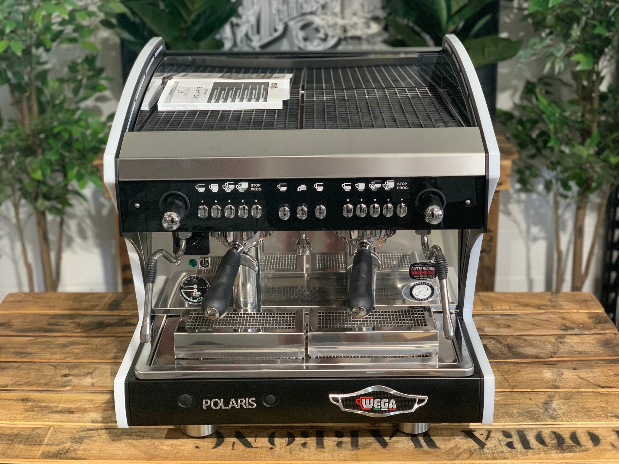 A new Wega Polaris Tron Compact 2 Group Espresso Coffee Machine with black finish  sitting on a wooden bench. There is greenery in the background. The image is taken from the front of the machine so that you can see the front of the machine, control panel, buttons and group heads.