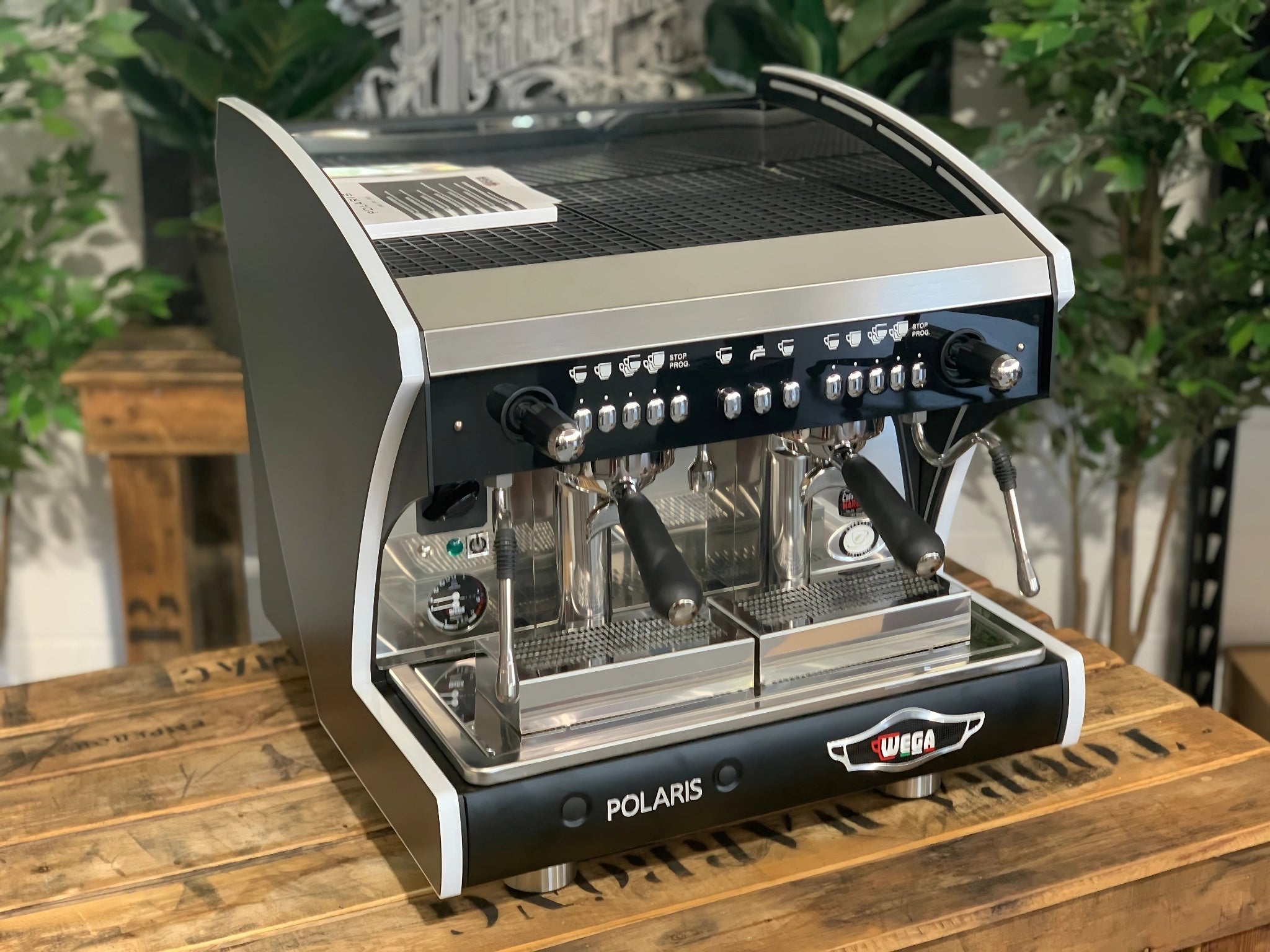 A new Wega Polaris Tron Compact 2 Group Espresso Coffee Machine with black finish is sitting on a wooden bench. There is greenery in the background. The image is taken from the front of the machine and the machine is sitting on an angle so that you can see the front of the machine and the side.