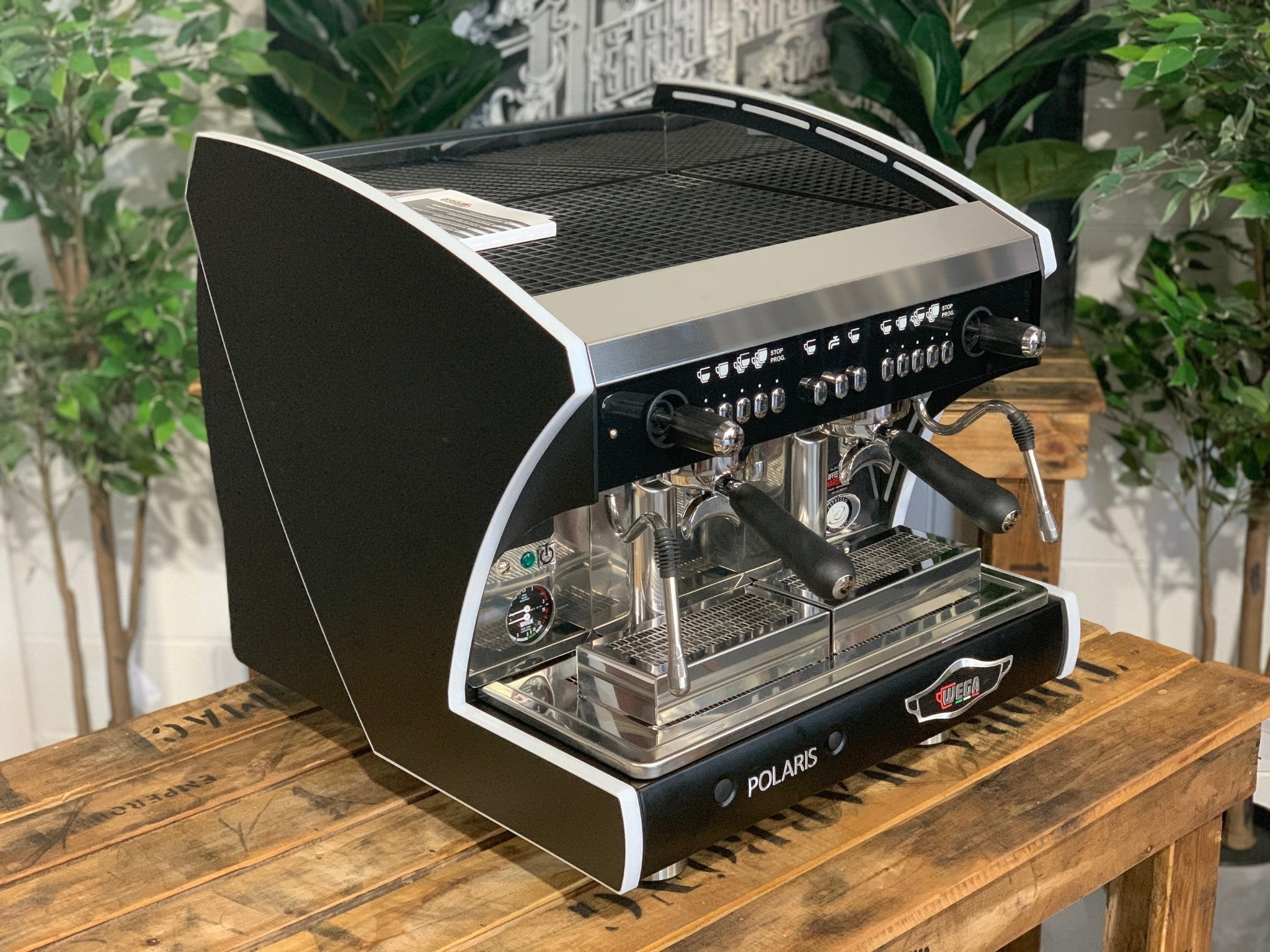 A new Wega Polaris Tron Compact 2 Group Espresso Coffee Machine with black finish  is sitting on a wooden bench. There is greenery in the background. The image is taken from the front of the machine and the machine is sitting on an angle so that you can see the front of the machine and the side.