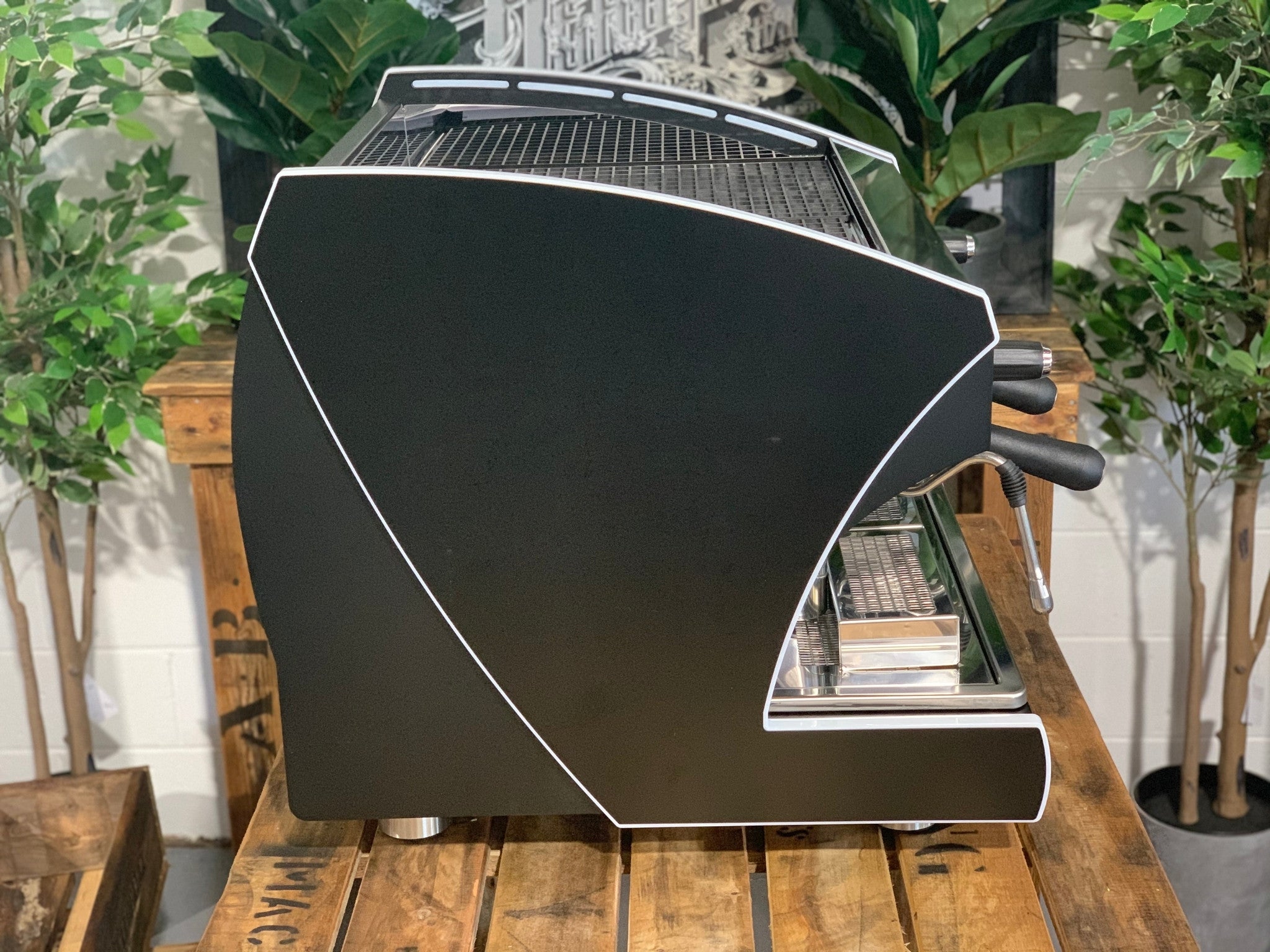 A new Wega Polaris Tron Compact 2 Group Espresso Coffee Machine with black finish sitting on a wooden bench. There is greenery in the background. The image is taken from the side of the machine.