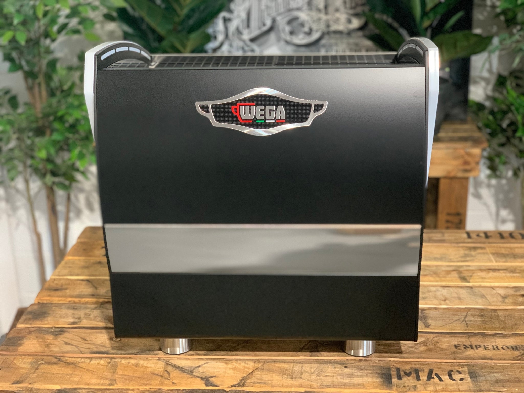 A new Wega Polaris Tron Compact 2 Group Espresso Coffee Machine with black finish is sitting on a wooden bench. There is greenery in the background. The image is taken from behind the machine so the back is visible. The Wega logo is situated in the middle close to the top of the back panel of the machine.