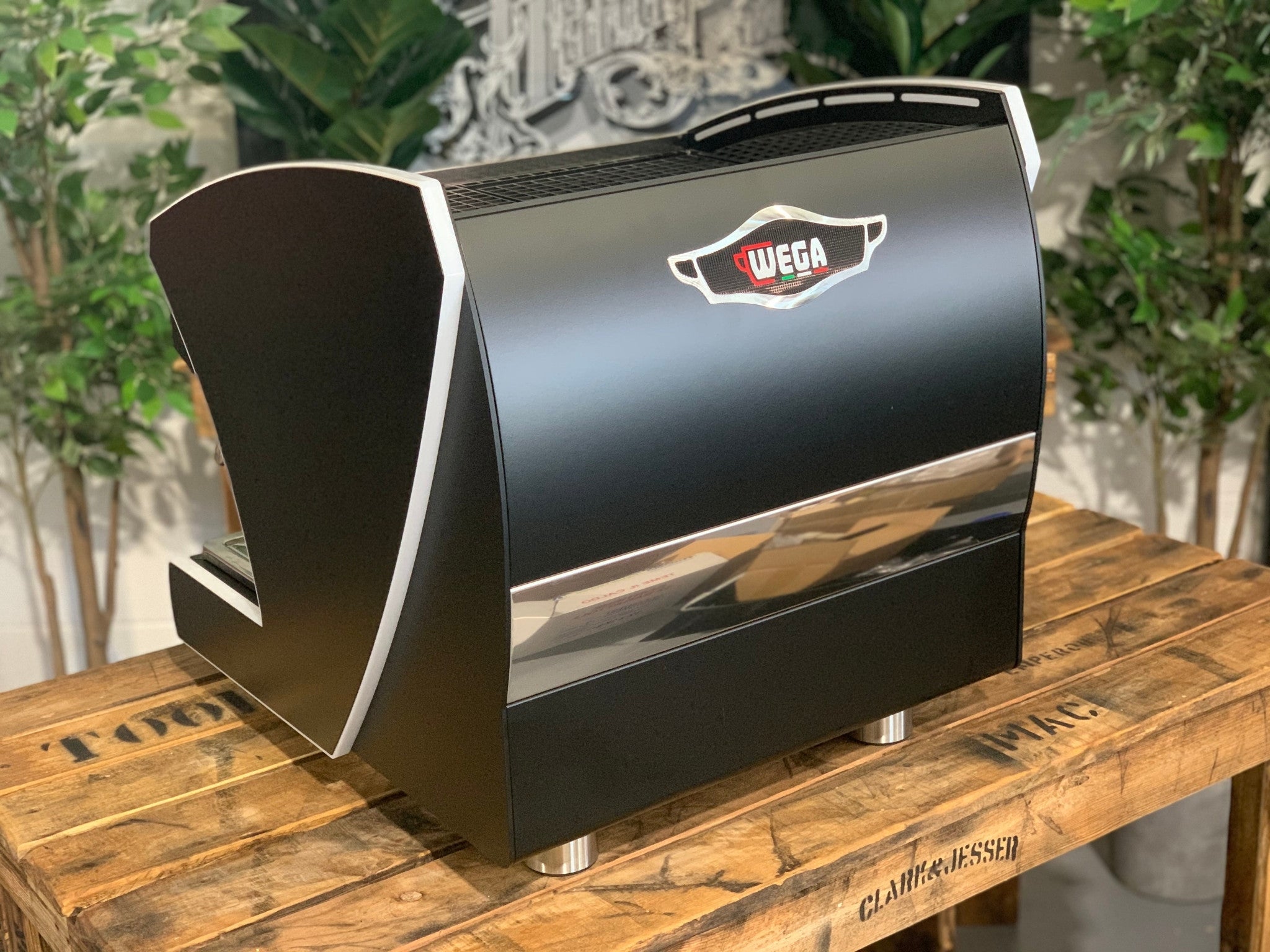 A new Wega Polaris Tron Compact 2 Group Espresso Coffee Machine with black finish sitting on a wooden bench. There is greenery in the background. The image is taken from behind the machine and the machine is sitting on an angle so that you can see the back of the machine and the side. The Wega logo is situated in the middle close to the top of the back panel of the machine.