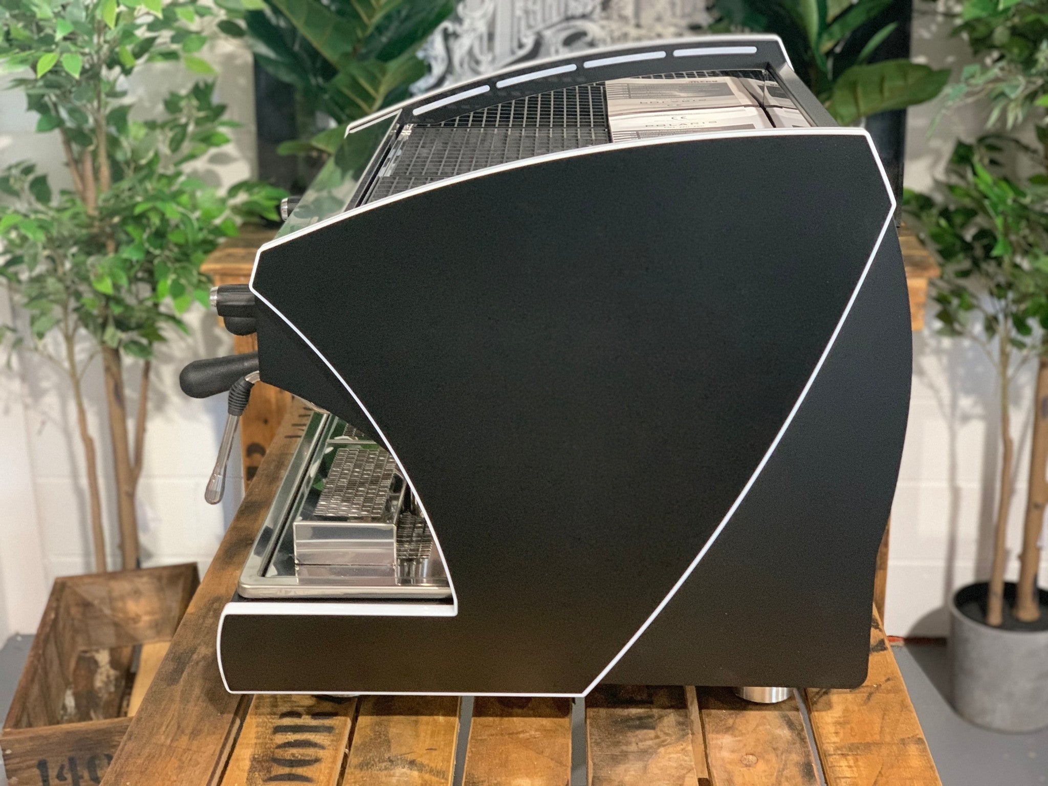 A new Wega Polaris Tron Compact 2 Group Espresso Coffee Machine with black finish sitting on a wooden bench. There is greenery in the background. The image is taken from the side of the machine.