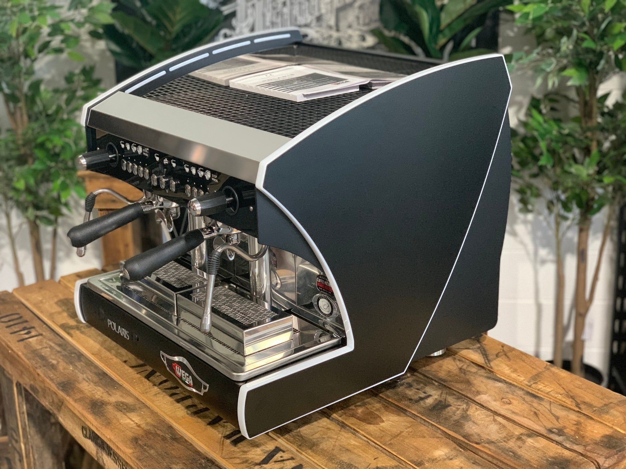 A new Wega Polaris Tron Compact 2 Group Espresso Coffee Machine with black finish  is sitting on a wooden bench. There is greenery in the background. The image is taken from the front of the machine and the machine is sitting on an angle so that you can see the front of the machine and the side.