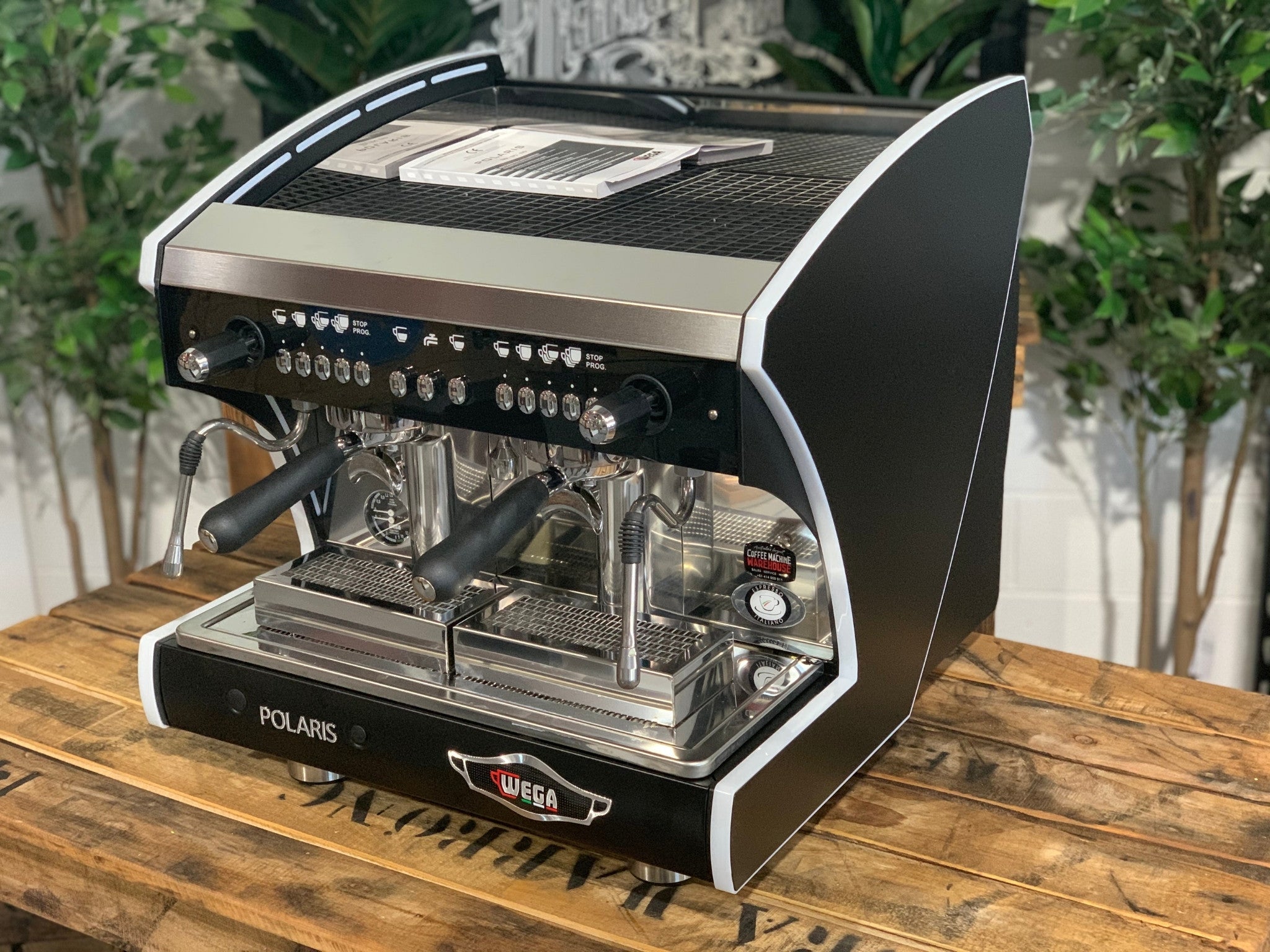 A new Wega Polaris Tron Compact 2 Group Espresso Coffee Machine with black finish  is sitting on a wooden bench. There is greenery in the background. The image is taken from the front of the machine and the machine is sitting on an angle so that you can see the front of the machine and the side.
