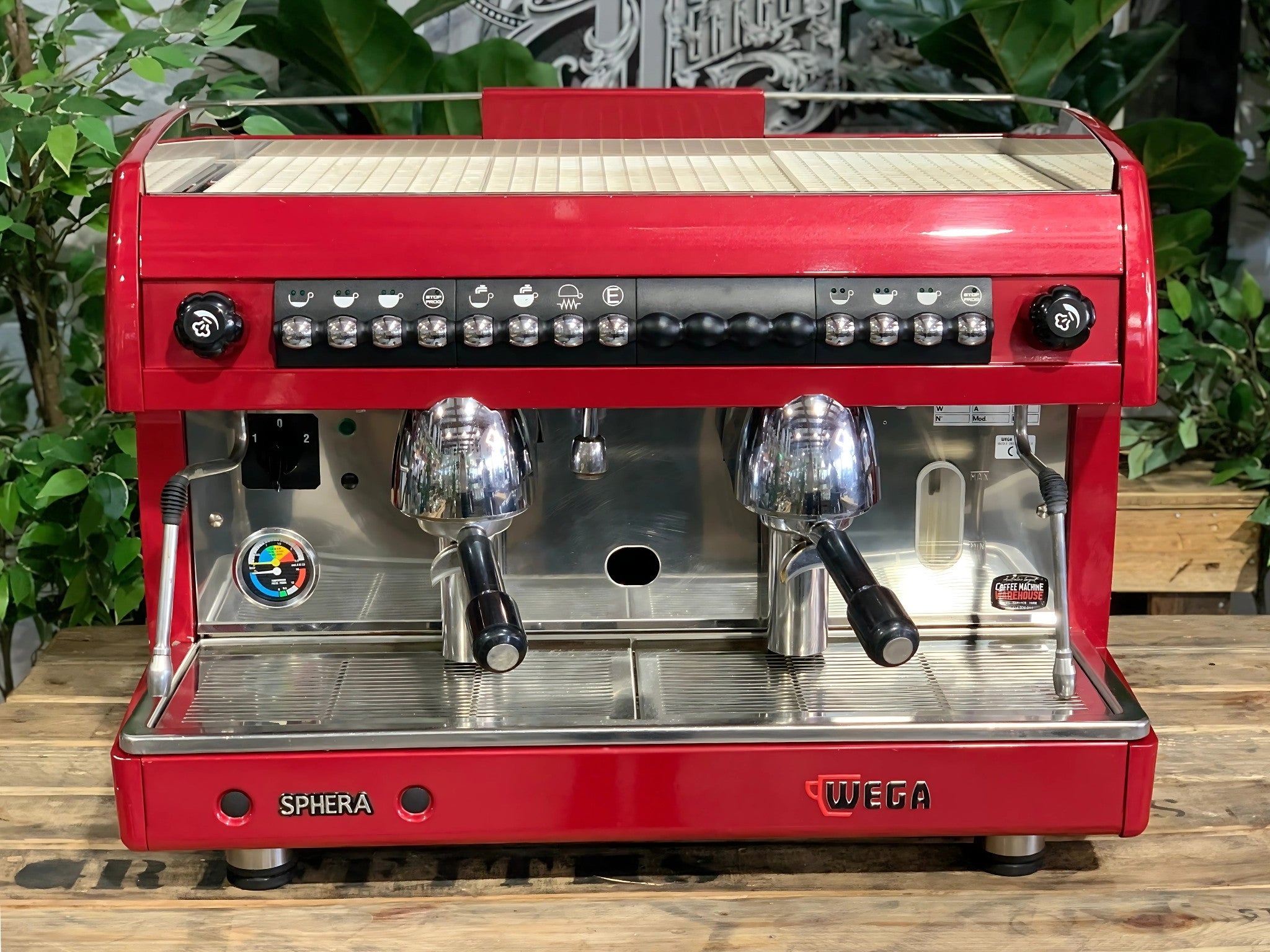 A Wega Sphera 2 Group Espresso Coffee Machine with Red finish sitting on a wooden bench. There is greenery in the background. The image is taken from the front of the machine so that you can see the front of the machine, control panel, buttons and group heads.