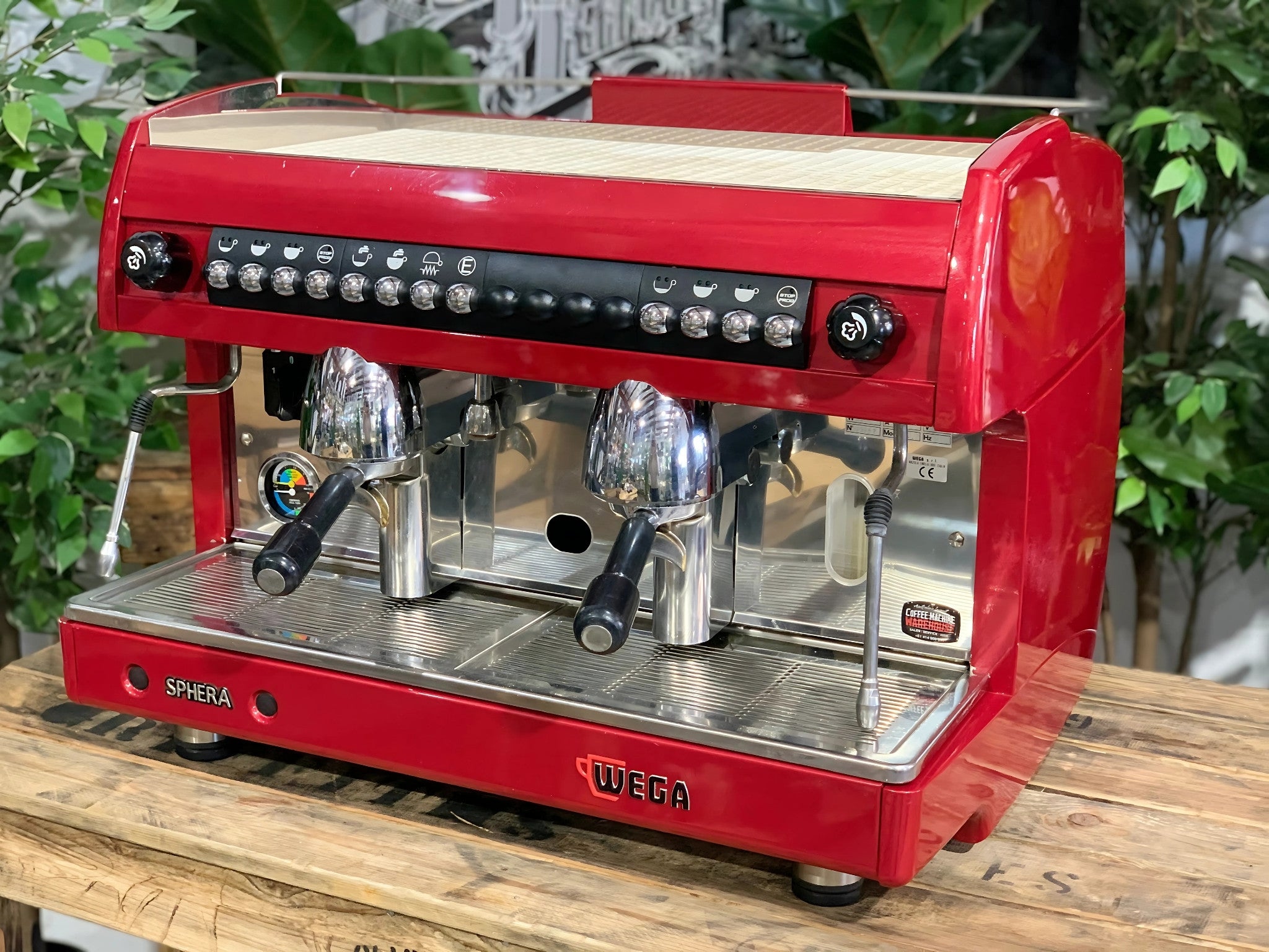 A Wega Sphera 2 Group Espresso Coffee Machine with Red finish is sitting on a wooden bench. There is greenery in the background. The image is taken from the front of the machine and the machine is sitting on an angle so that you can see the front of the machine and the side.