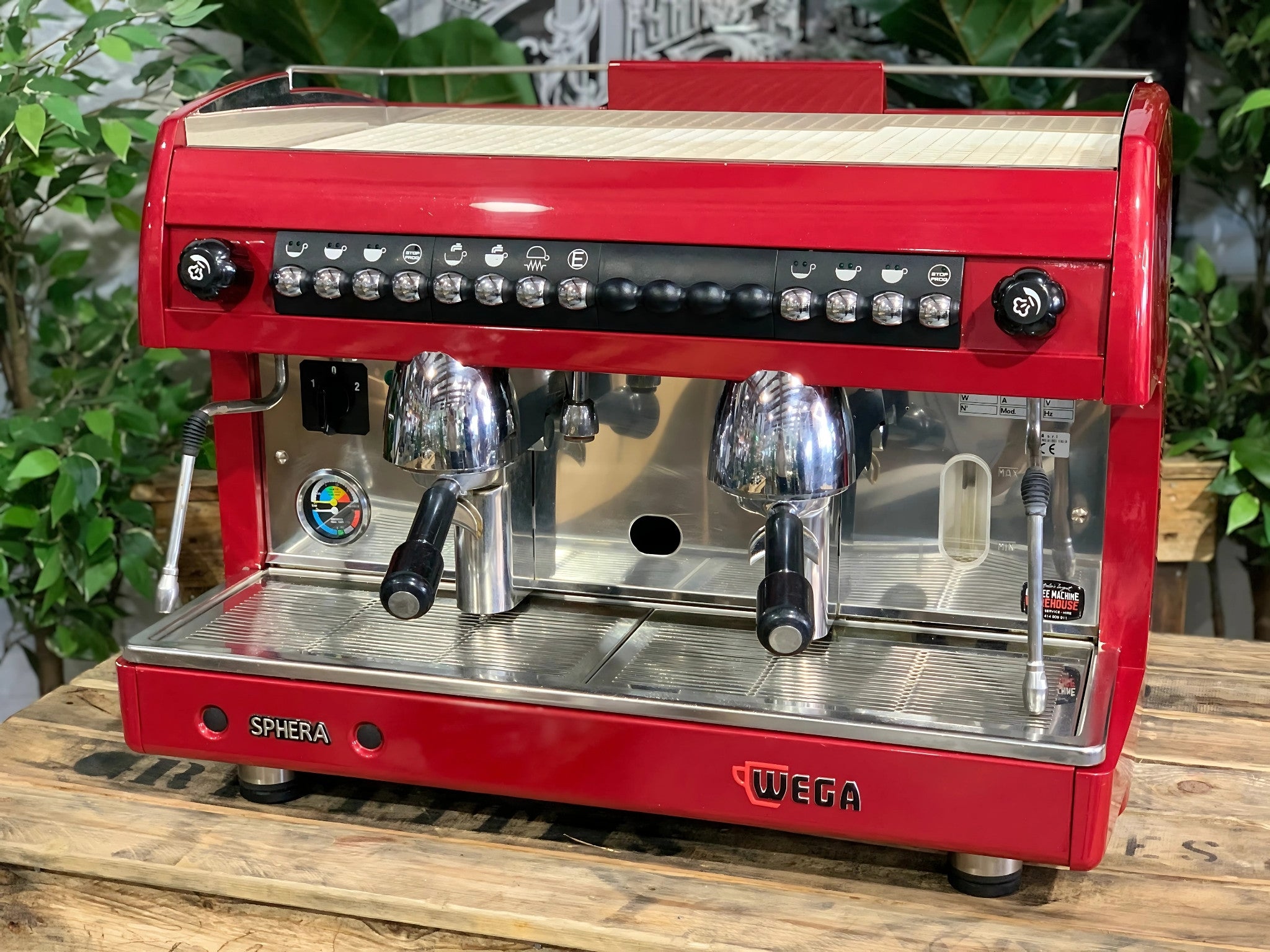 A Wega Sphera 2 Group Espresso Coffee Machine with Red finish sitting on a wooden bench. There is greenery in the background. The image is taken from the front of the machine so that you can see the front of the machine, control panel, buttons and group heads.