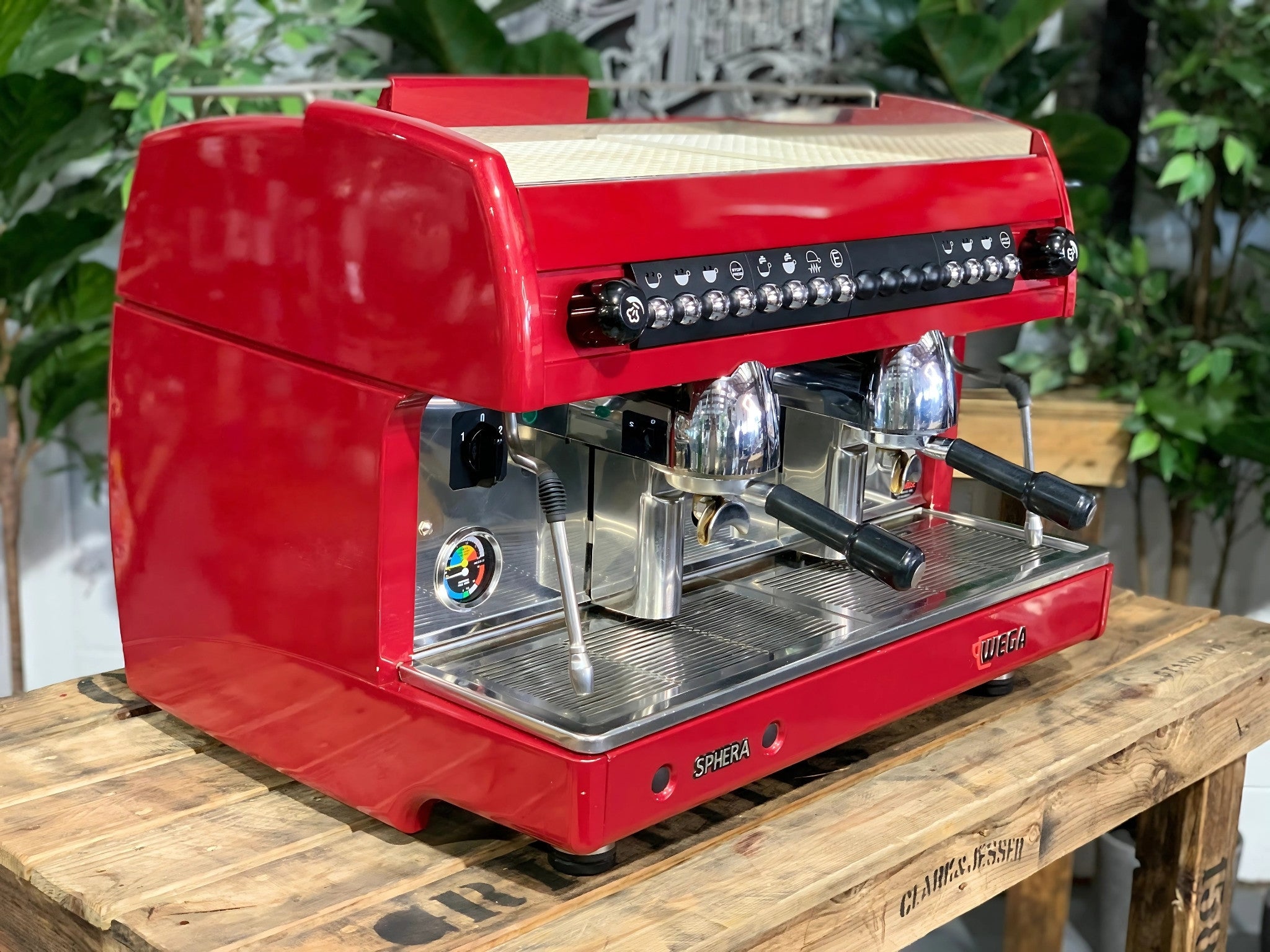A Wega Sphera 2 Group Espresso Coffee Machine with Red finish is sitting on a wooden bench. There is greenery in the background. The image is taken from the front of the machine and the machine is sitting on an angle so that you can see the front of the machine and the side.