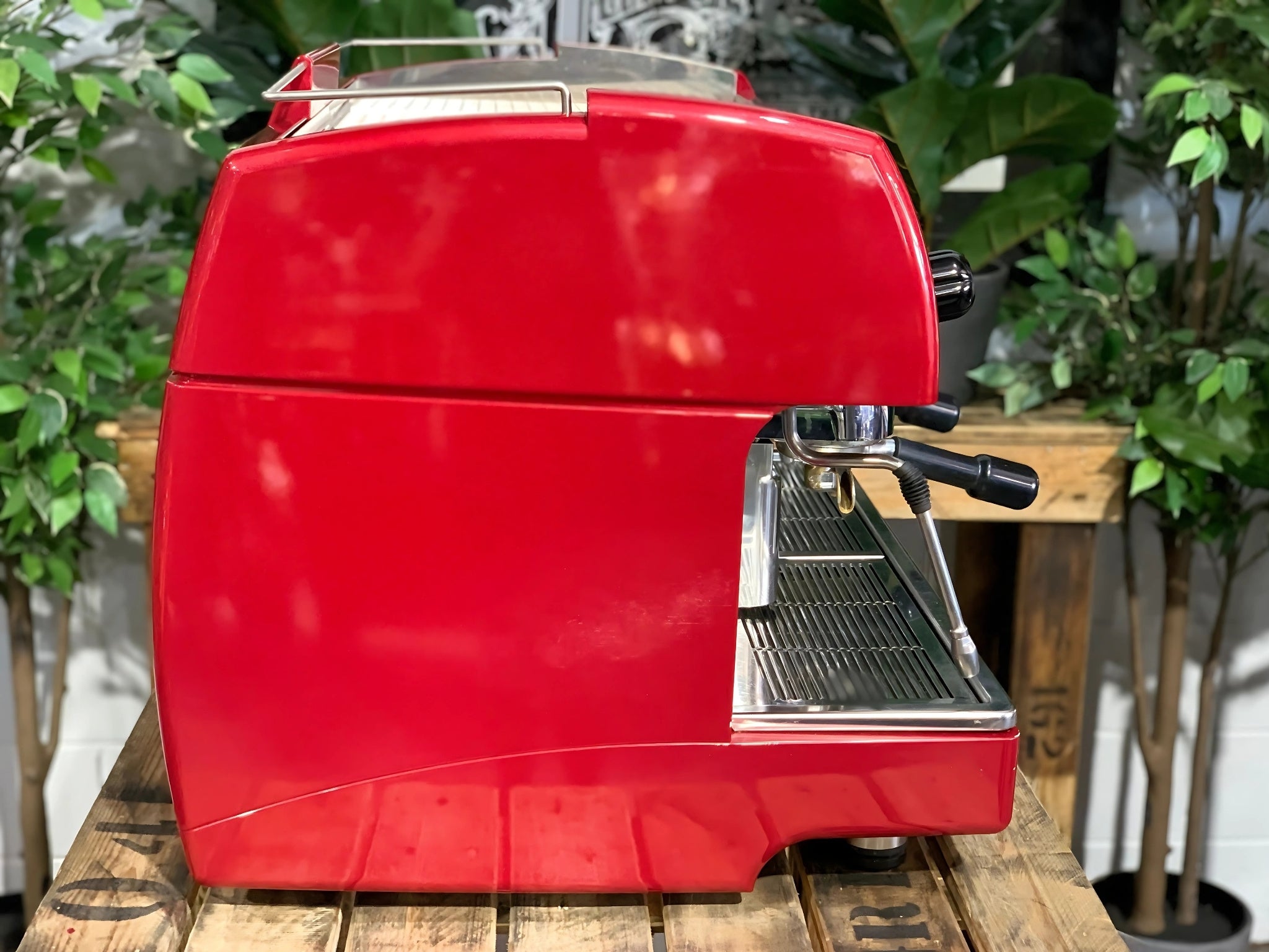 A Wega Sphera 2 Group Espresso Coffee Machine with Red finish sitting on a wooden bench. There is greenery in the background. The image is taken from the side of the machine.