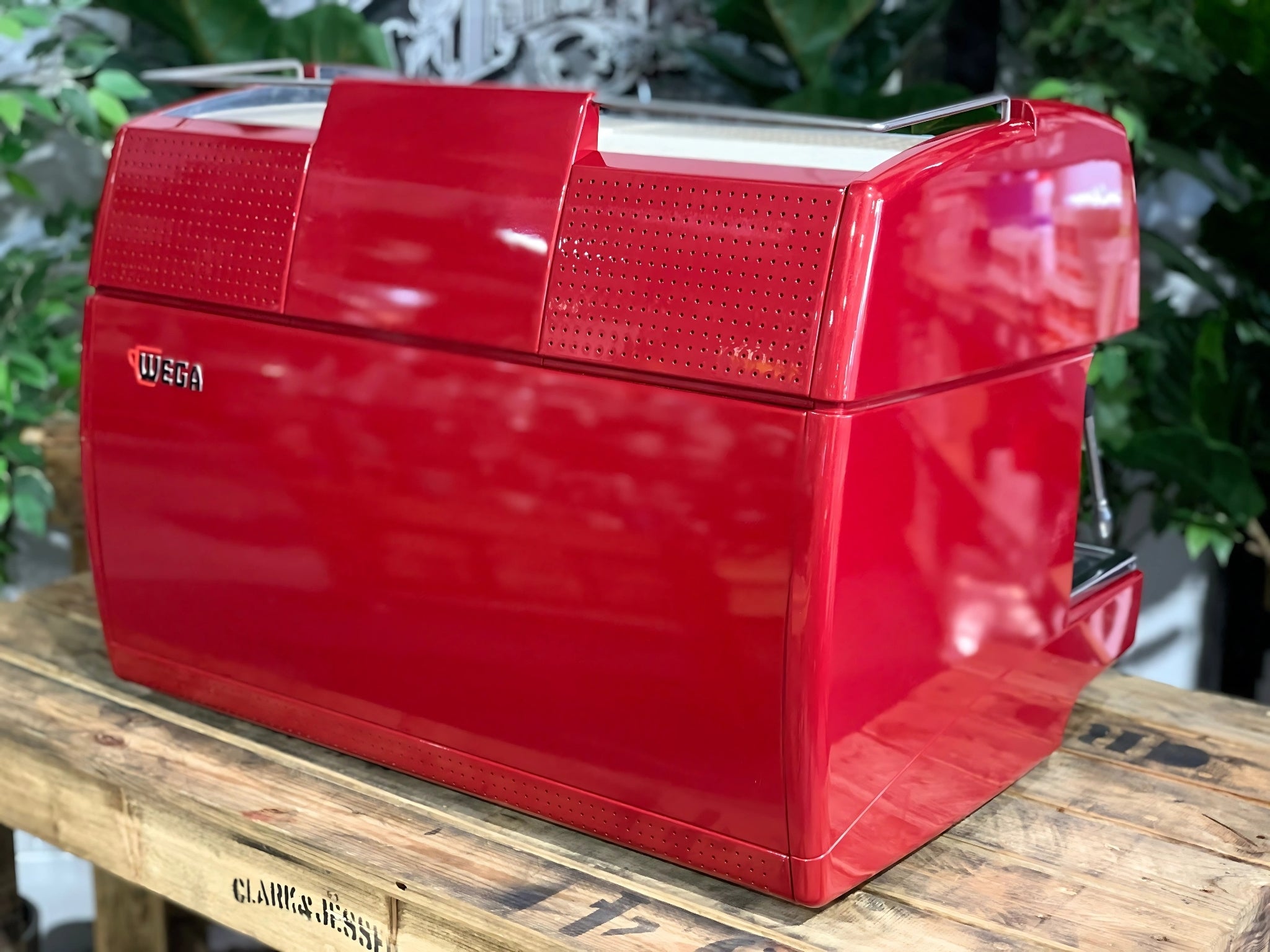 A Wega Sphera 2 Group Espresso Coffee Machine with Red finish sitting on a wooden bench. There is greenery in the background. The image is taken from behind the machine and the machine is sitting on an angle so that you can see the back of the machine and the side.