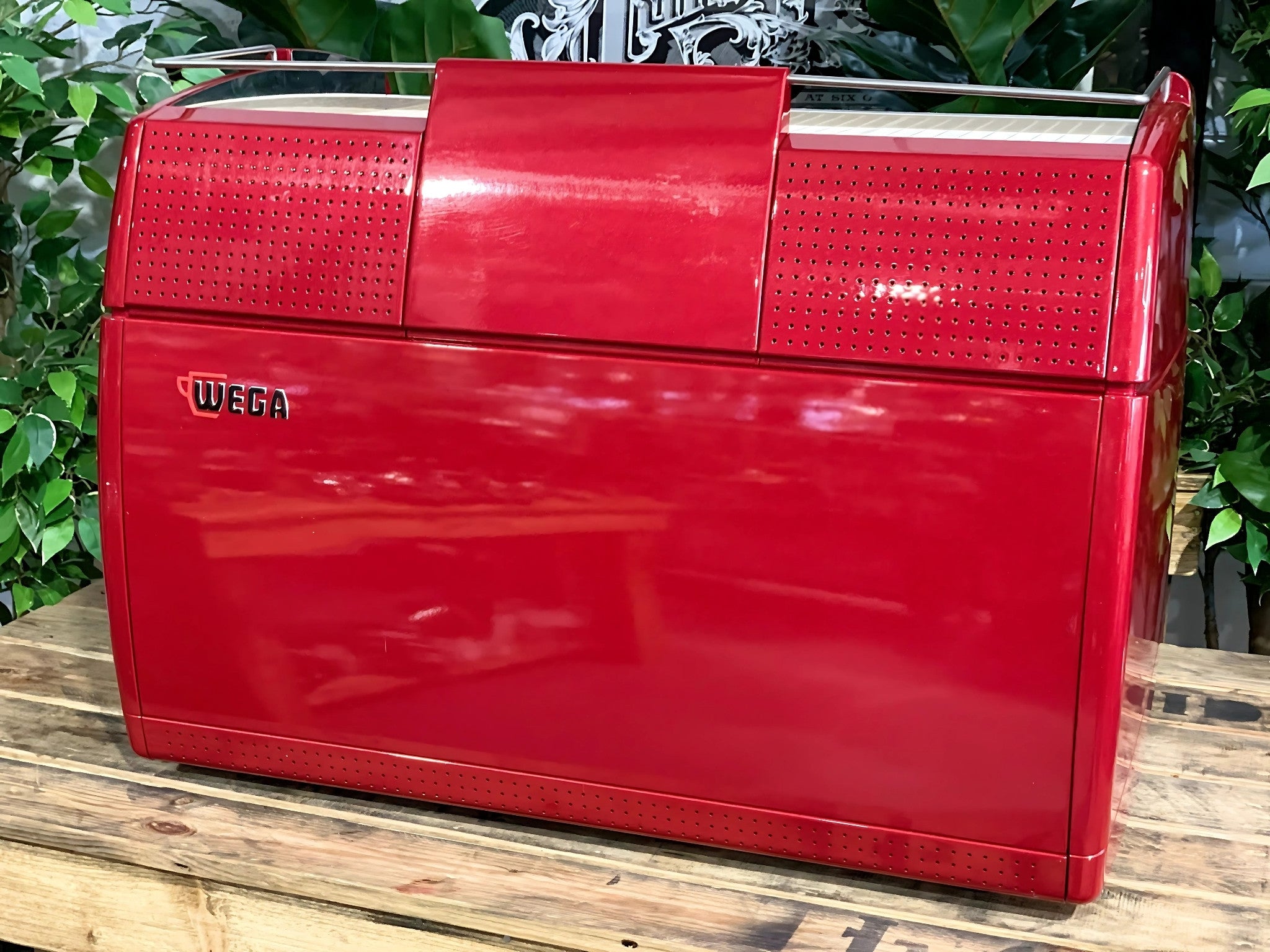 A Wega Sphera 2 Group Espresso Coffee Machine with Red finish is sitting on a wooden bench. There is greenery in the background. The image is taken from behind the machine so the back is visible. The machine is red and the Wega logo is on the back of the machine placed in the middle of the left hand side.