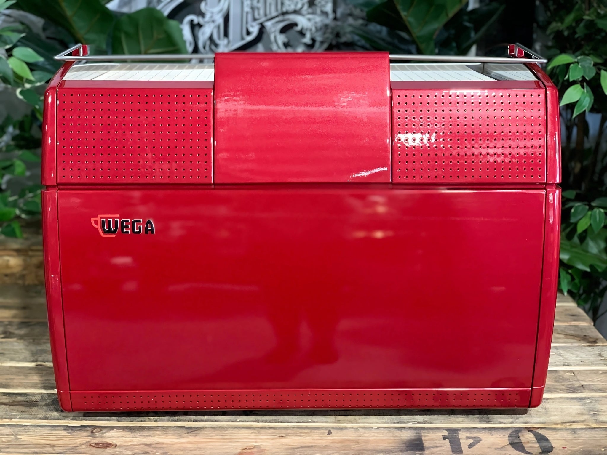 A Wega Sphera 2 Group Espresso Coffee Machine with Red finish is sitting on a wooden bench. There is greenery in the background. The image is taken from behind the machine so the back is visible. The machine is red and the Wega logo is on the back of the machine placed in the middle of the left hand side.